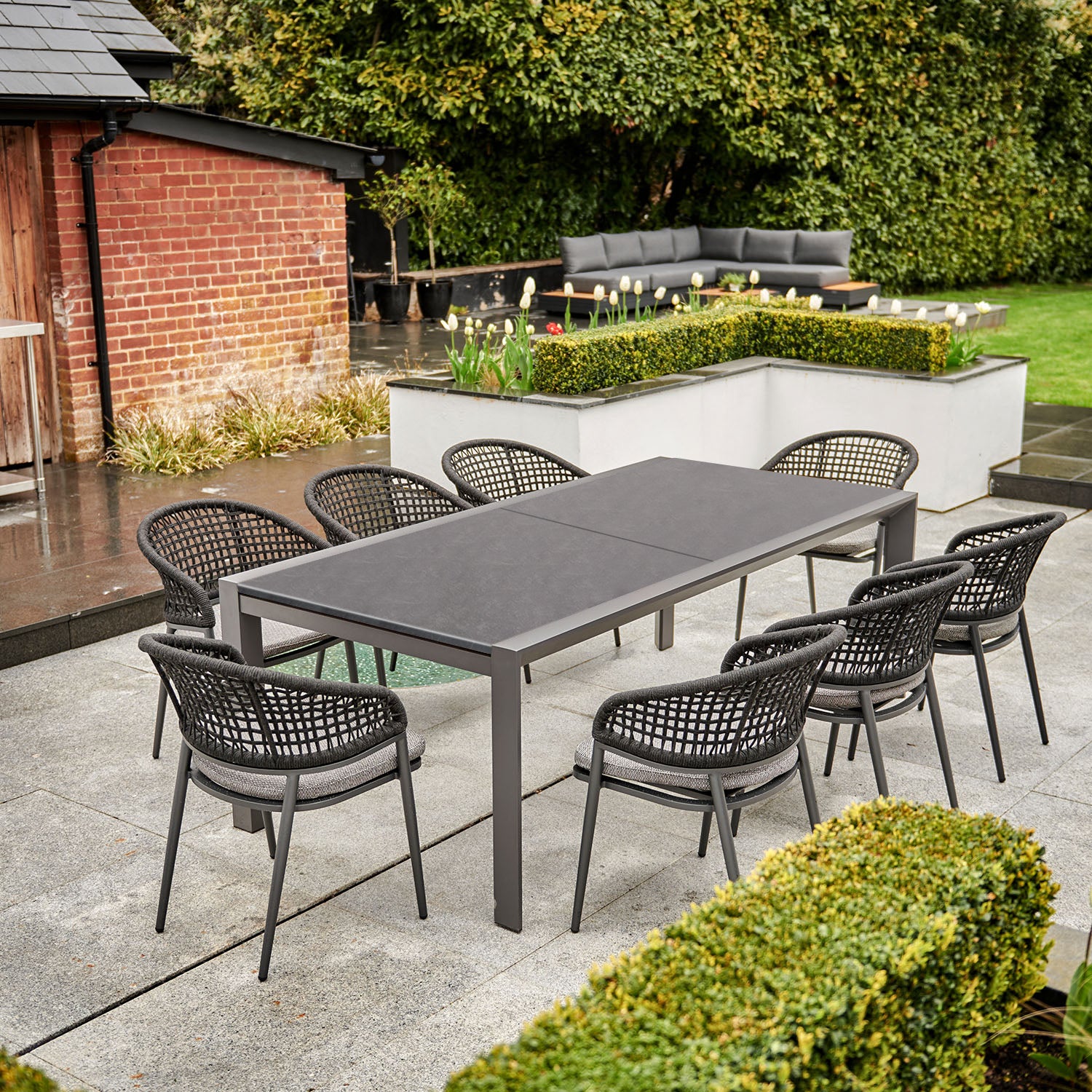 Outdoor patio with a Kalama 10 Seat Rectangular Extending Dining Set with Ceramic Table in Charcoal, featuring eight weather-proof aluminum wicker chairs, and a nearby seating area with a sofa, all set in a lush garden.