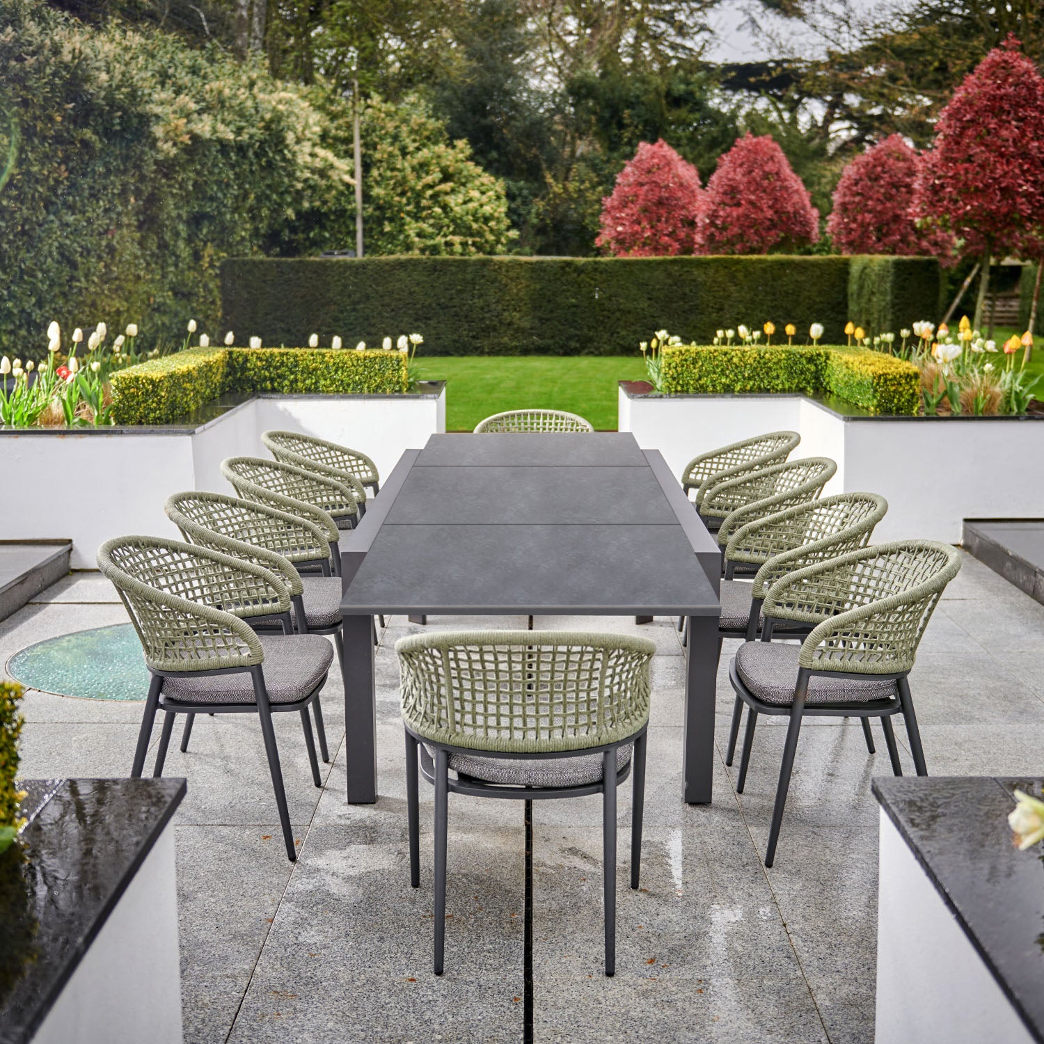 Outdoor dining area with the Kalama 10 Seat Rectangular Extending Dining Set, featuring a ceramic table in olive green and ten woven chairs, surrounded by greenery and red shrubs.