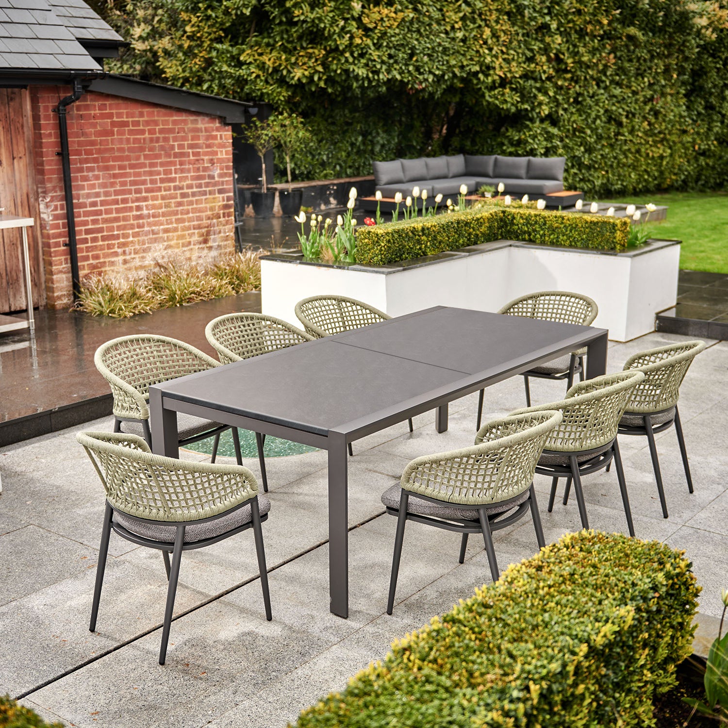 Outdoor dining area adorned with the Kalama 10 Seat Rectangular Extending Dining Set, featuring a ceramic table in olive green and eight woven chairs on a patio, surrounded by lush greenery and a garden bed.