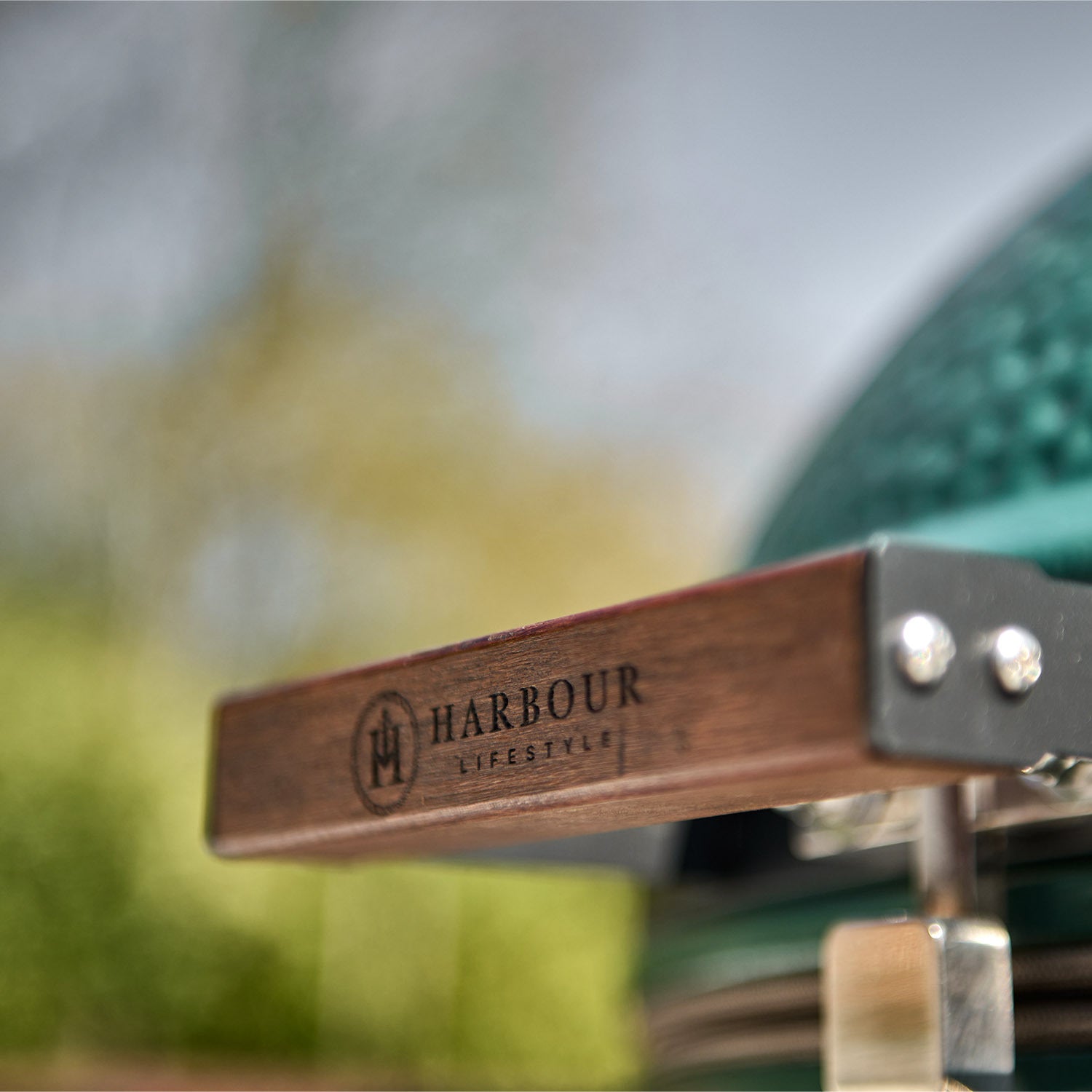 Close-up of the wooden handle on a Kamado BBQ 22" Ceramic Grill in Matte Green labeled "Harbour Lifestyle," with a blurred background.