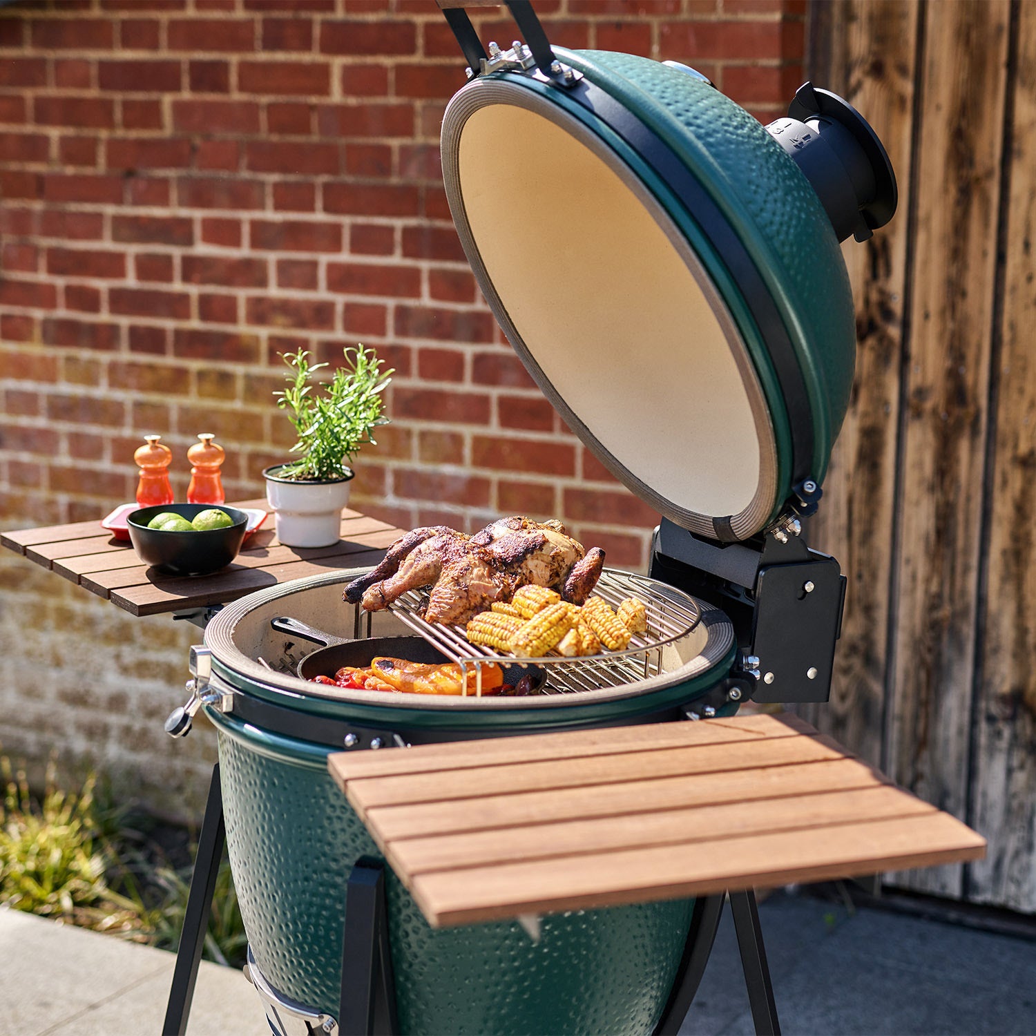 Matte Green Kamado BBQ 22" Ceramic Grill with open lid, cooking assorted meats and vegetables outdoors next to a plant and condiments.