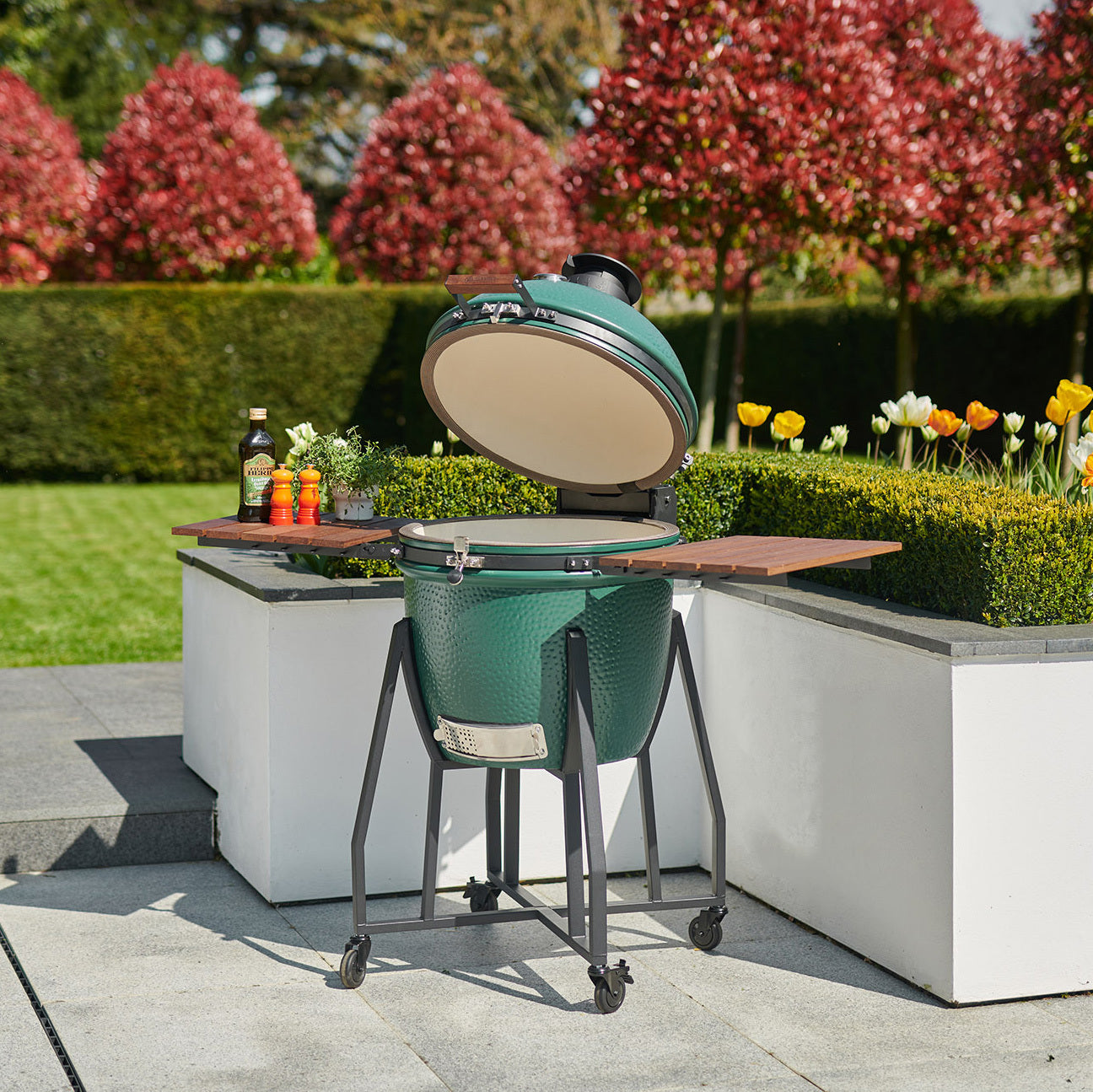 A Kamado BBQ 22" Ceramic Grill in Matte Green, with its lid open, stands on a patio near a garden filled with tulips and trimmed hedges.