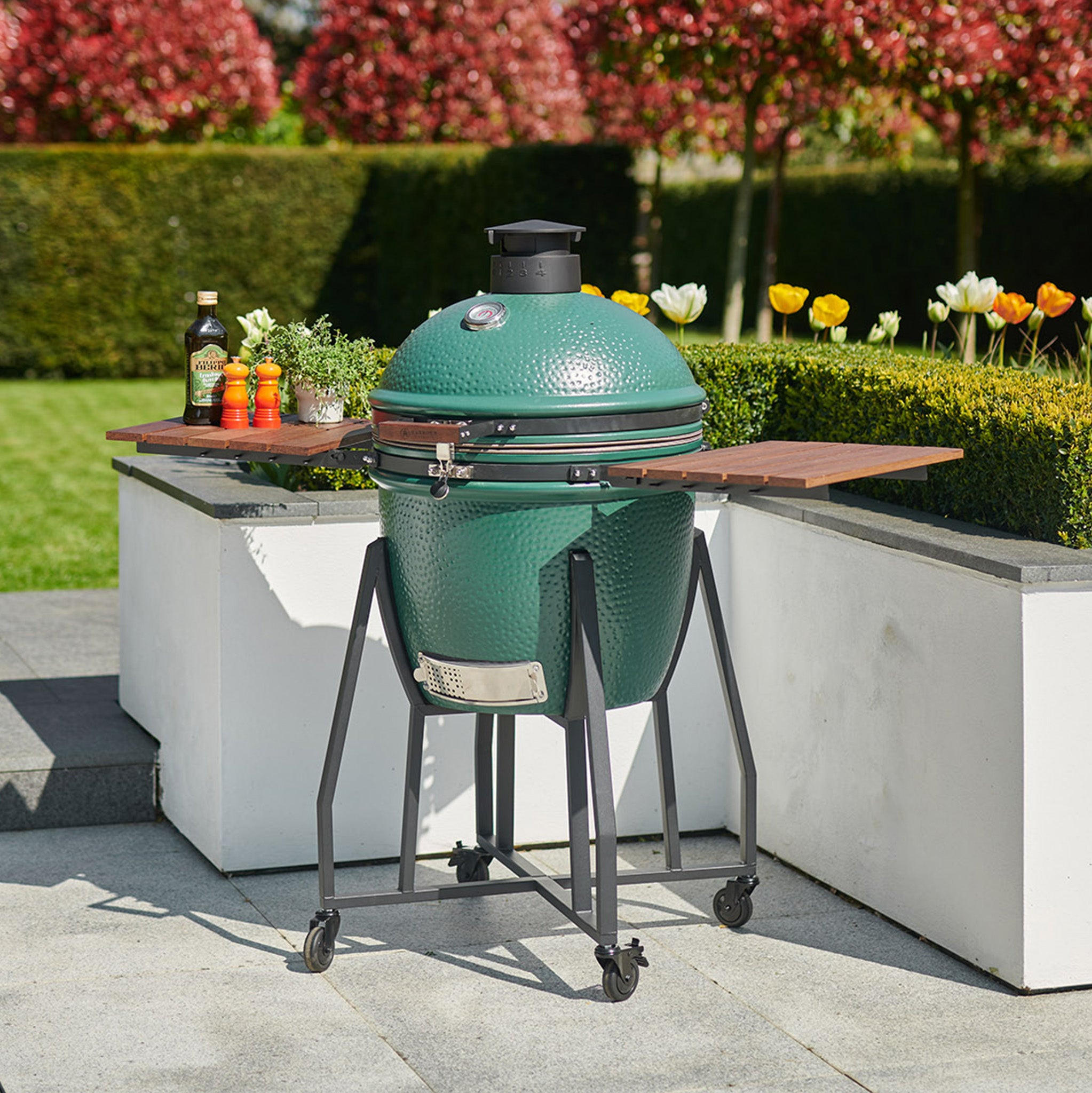 A Kamado BBQ 22" Ceramic Grill in Matte Green, complete with wooden side tables, sits on a patio near a garden filled with colorful flowers and hedges.