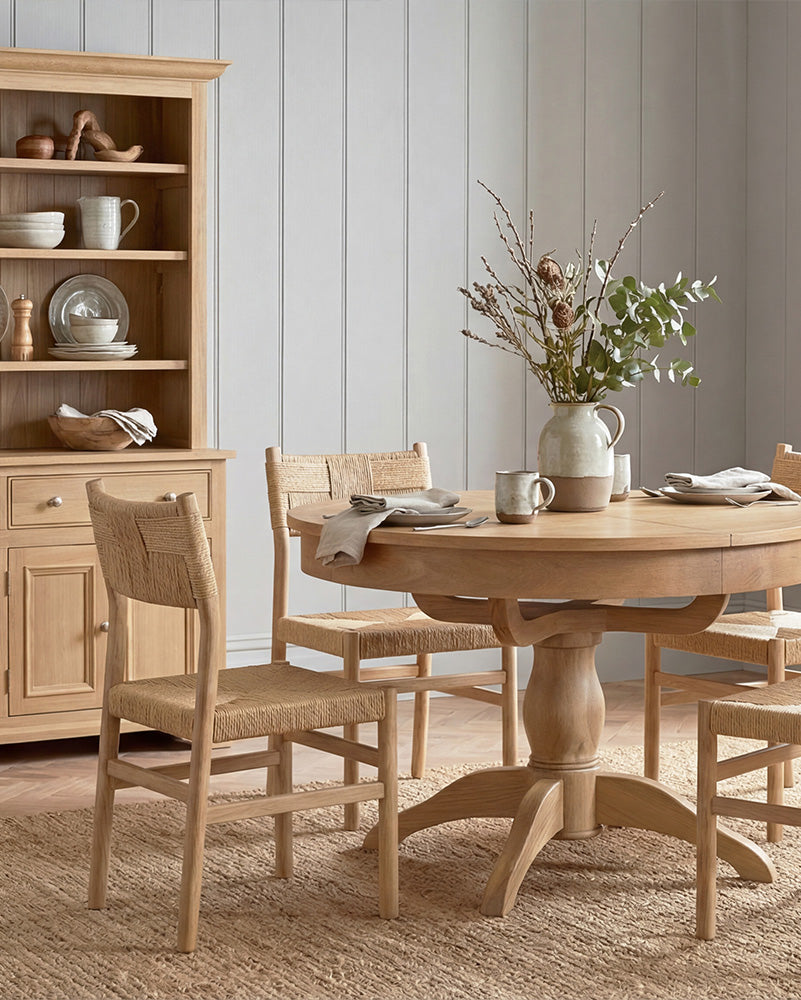 An Amberley wooden dining table with 4 chairs in a room with wooden flooring and a bookshelf.