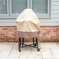 The Rain Cover for 22" Kamado Grills, crafted from premium waterproof fabric, protects the grill on wheels positioned by a brick wall and window blinds on a tiled patio.