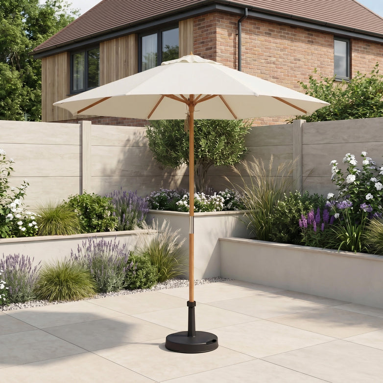 The Kratos 2.5m Round Wooden Parasol in Beige stands in a landscaped garden, providing UV protection among blooming flowers, with a brick house visible in the background.