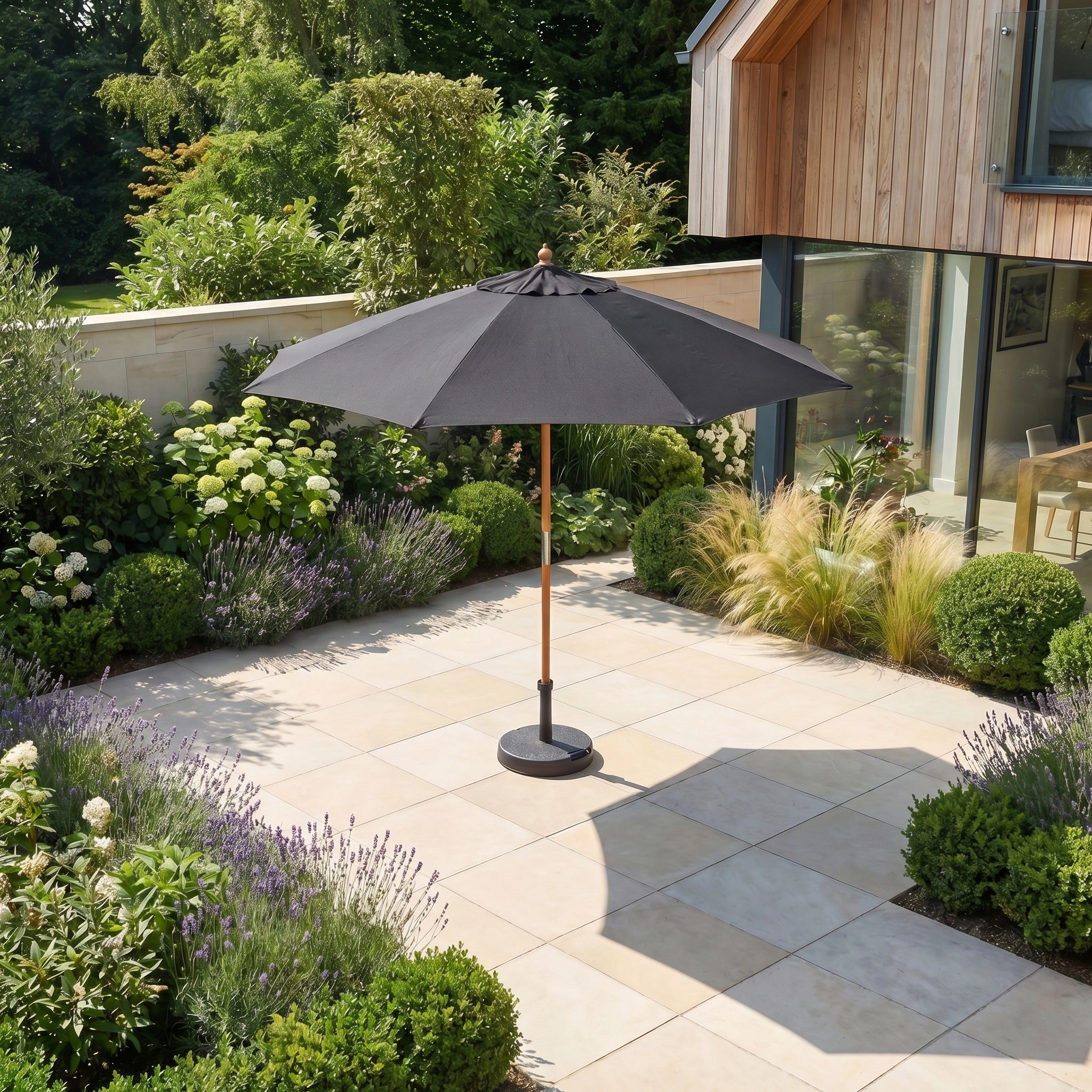A Kratos 2.5m Round Wooden Parasol in Black with a UV rating of 50+ stands on a tiled terrace, surrounded by lush greenery and flowering plants.