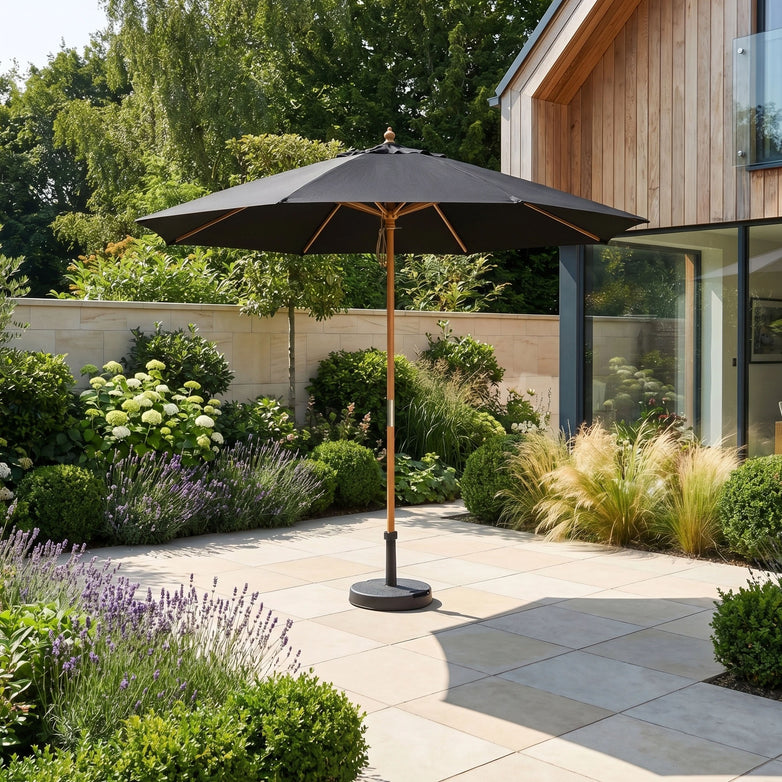 The Kratos 2.5m Round Wooden Parasol in Black stands on a tiled terrace by a modern house, surrounded by lush garden plants, providing stylish UV sun protection.