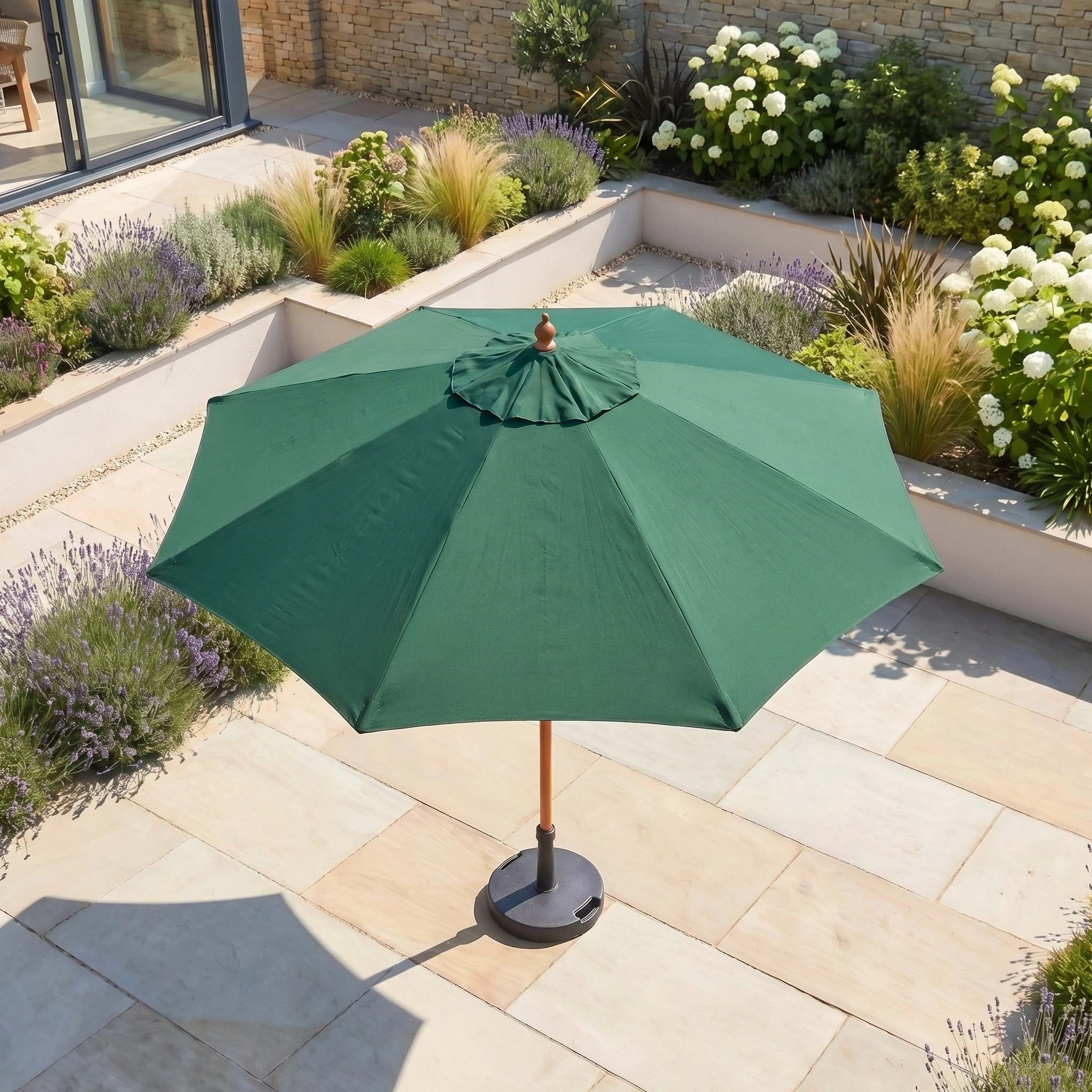 The Kratos 2.5m Round Wooden Parasol in Green stands open on a stone-tiled terrace amidst landscaped gardens, providing stylish UV protection.