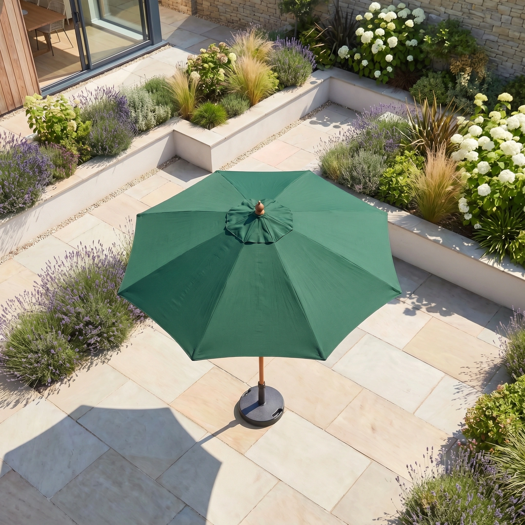 The Kratos 2.5m Round Wooden Parasol in Green stands in a landscaped garden with stone tiles and raised flower beds, adding style and UV protection.