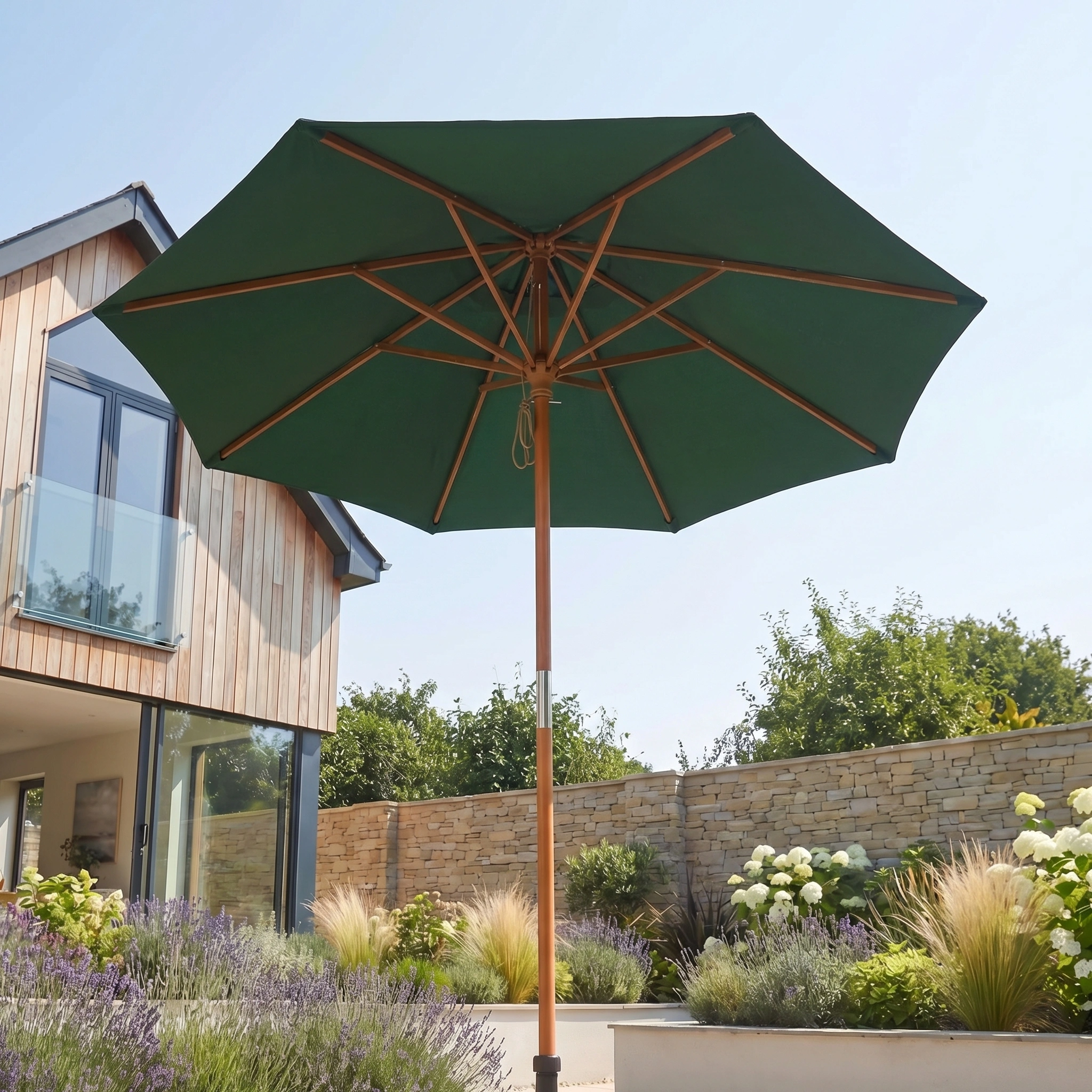 The Kratos 2.5m Round Wooden Parasol in Green provides UV protection as it shades a sunny garden with flowers, positioned near a modern house with large windows.