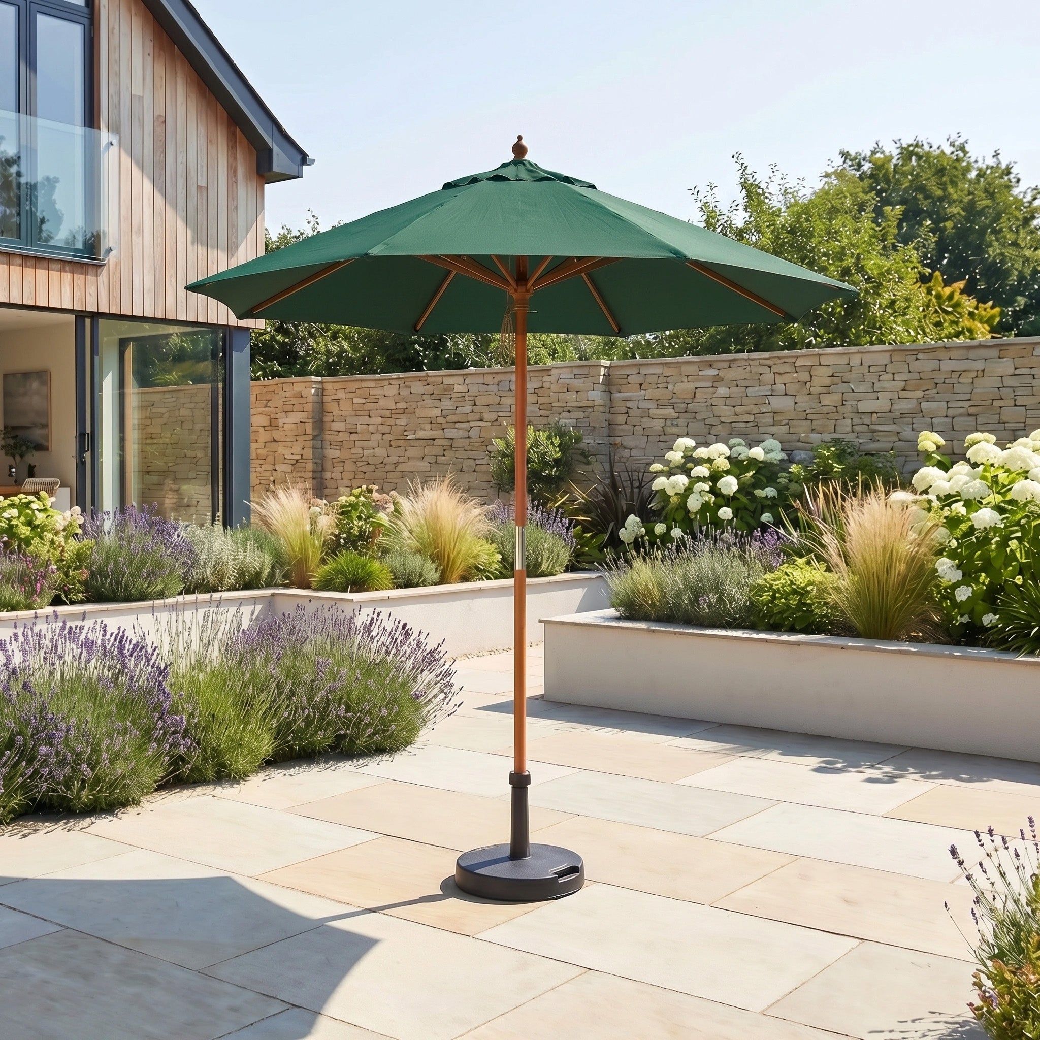 The Kratos 2.5m Round Wooden Parasol in Green offers UV protection as it stands on a sunny stone terrace, surrounded by blooming flowers and tall grass in the garden.