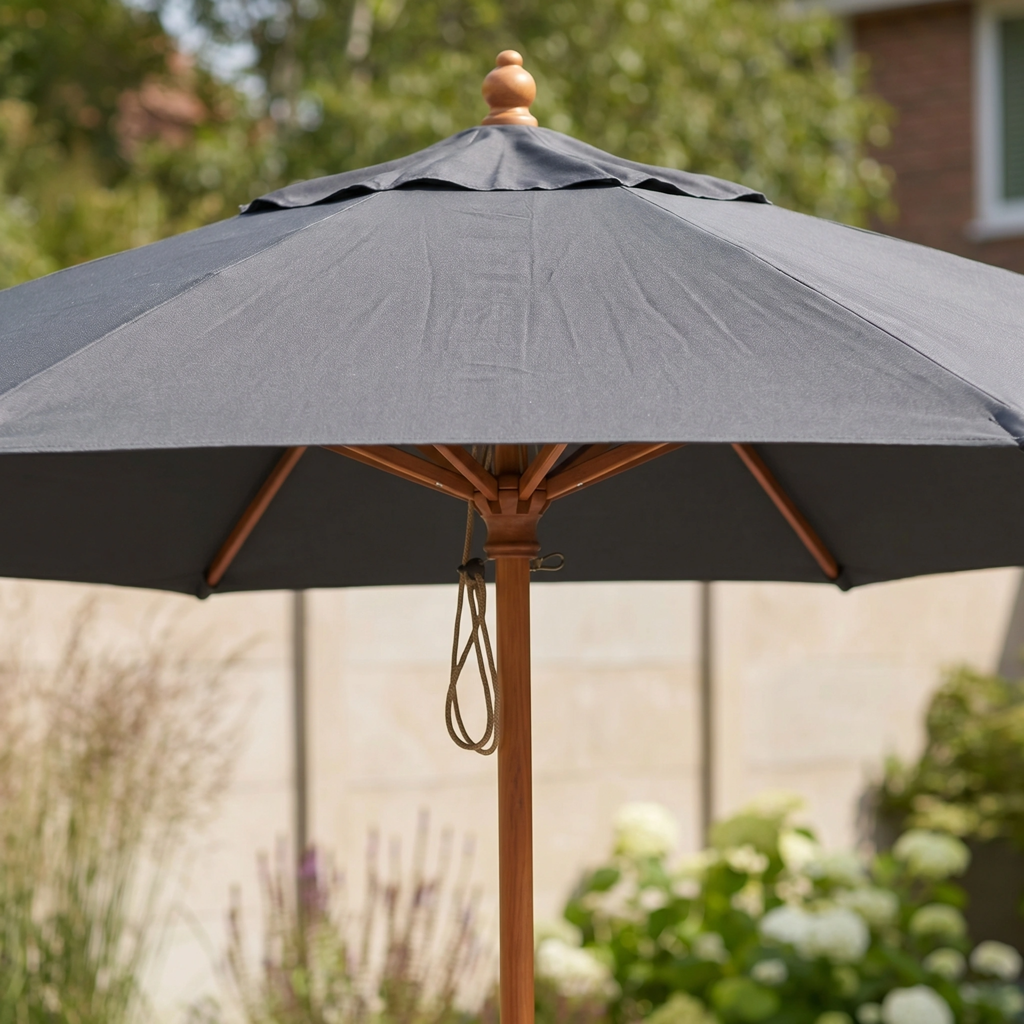 The Kratos 2.5m Round Wooden Parasol in Grey stands in a sunny garden, featuring a sturdy pole and offering UV 50+ sun protection amid lush greenery.