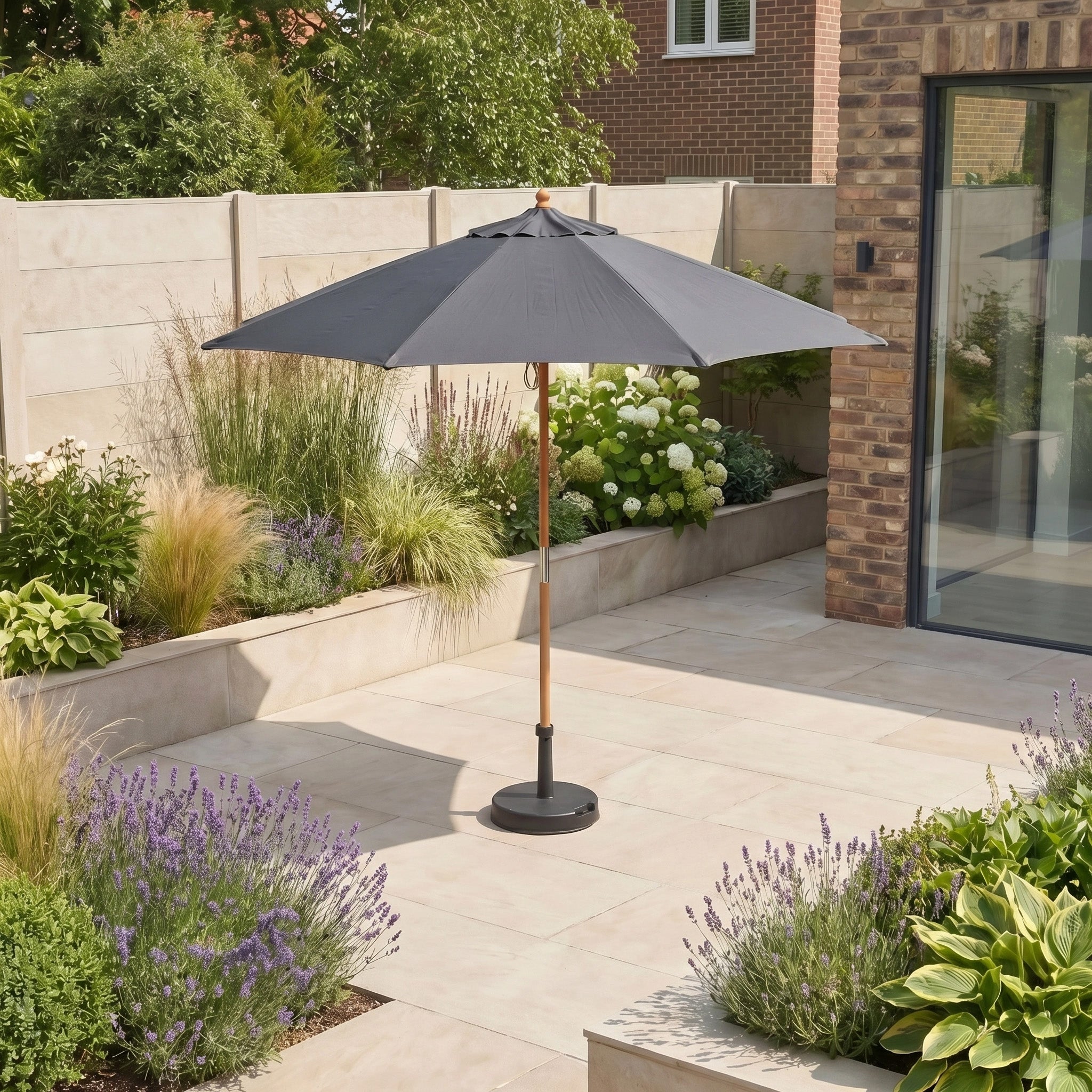 The Kratos 2.5m Round Wooden Parasol in Grey, with UV50+ protection, stands on a tiled terrace surrounded by plants and flowers in a modern garden.