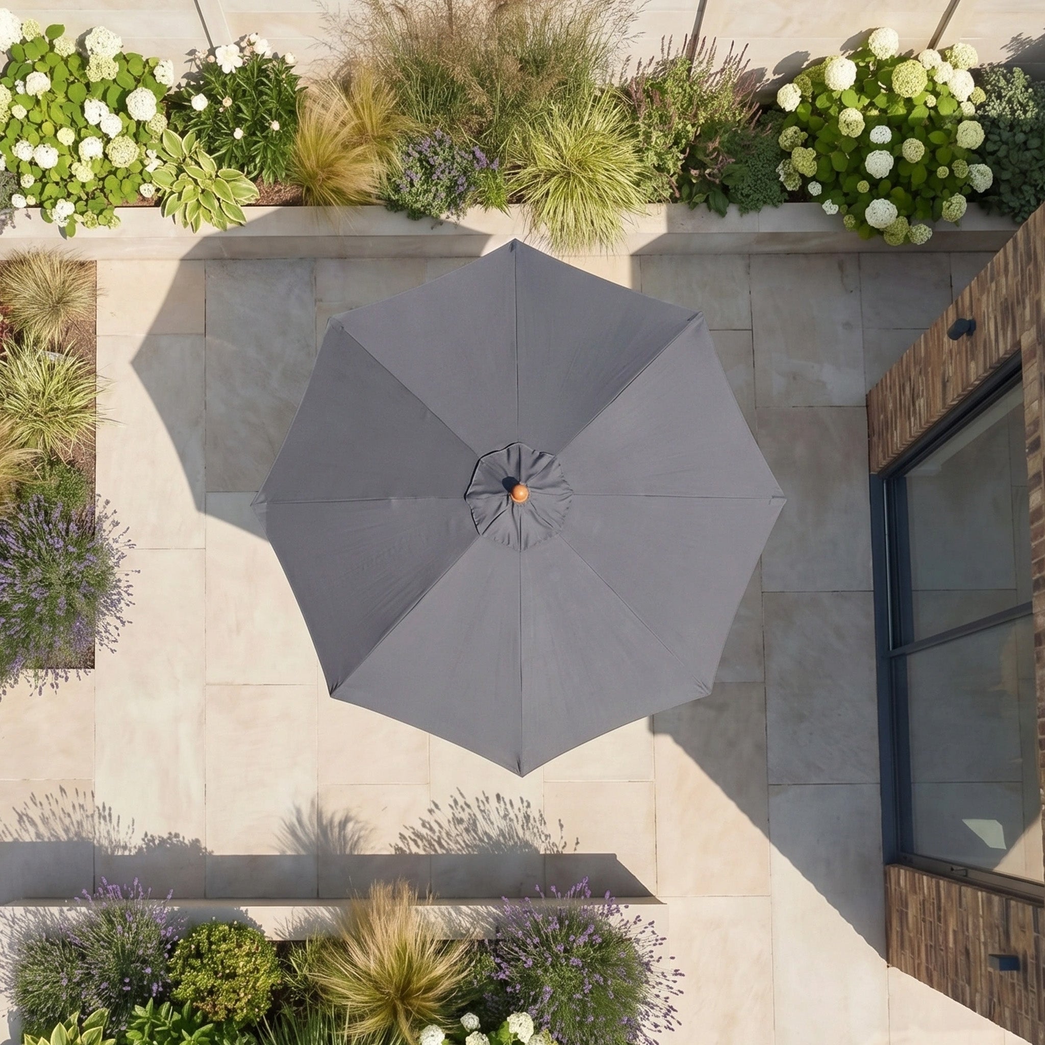 Aerial view of the Kratos 2.5m Round Wooden Parasol in Grey (UV 50+), serving as a sun protector, surrounded by plants and flowers on a stone terrace.