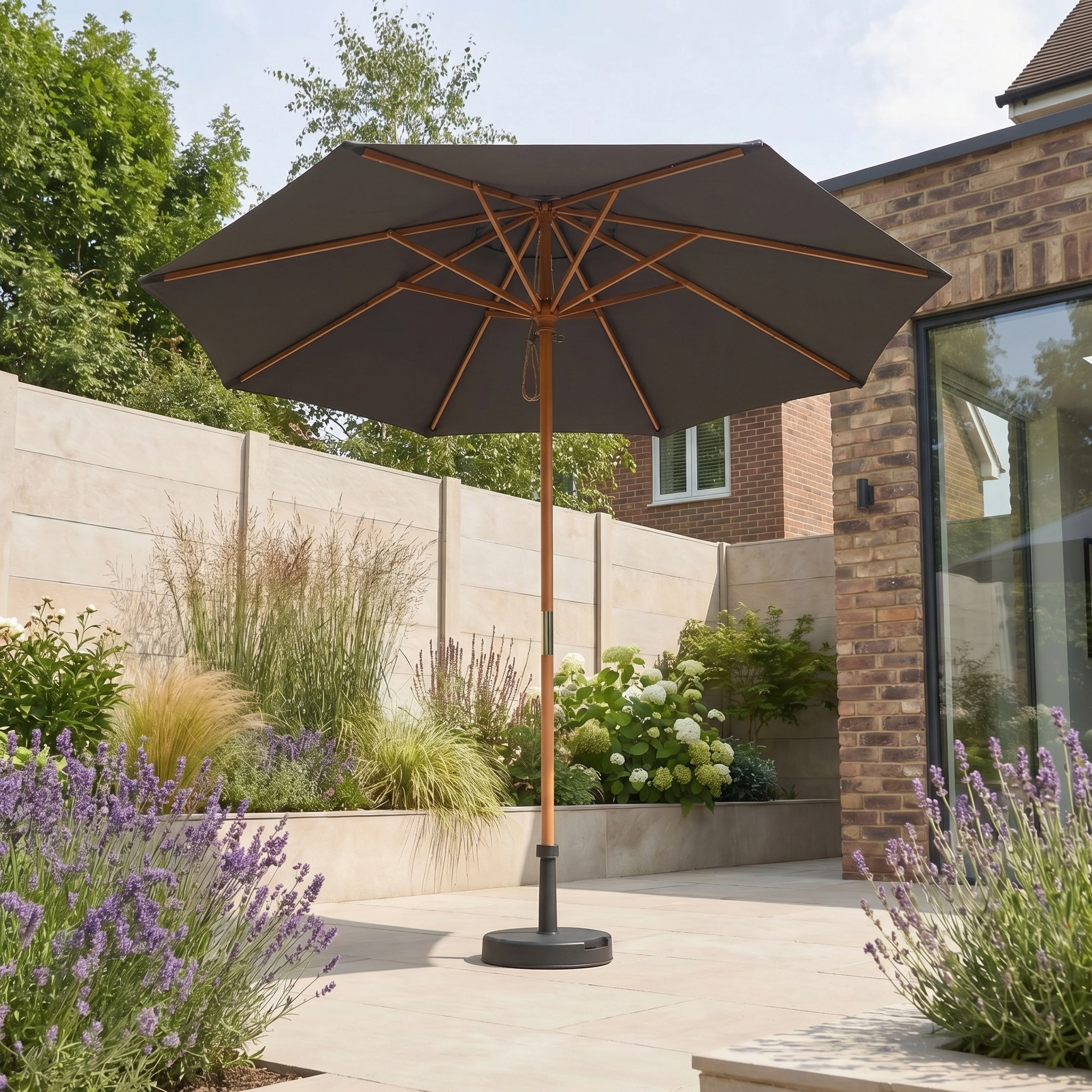 The Kratos 2.5m Round Wooden Parasol in Grey, UV 50+ rated, stands in a landscaped backyard with lush plants and a brick house, offering stylish and effective sun protection.