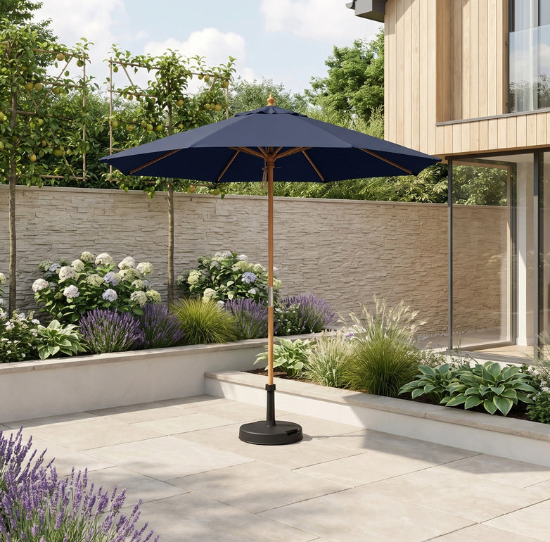 The Kratos 2.5m Round Wooden Parasol in Navy offers stylish shade and UV protection on a sunny stone terrace, surrounded by green plants and blooming flowers.