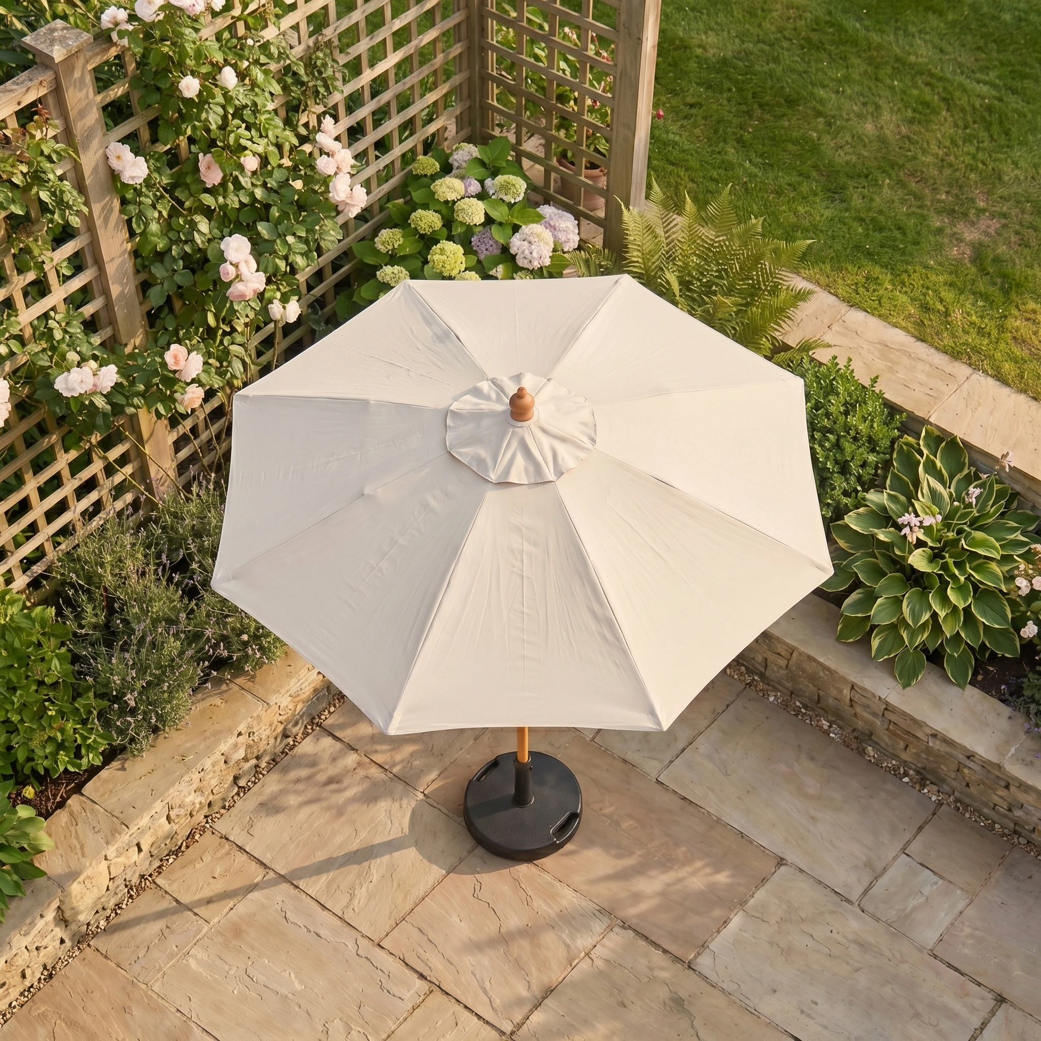 The Kratos 2m Round Wooden Parasol in Beige stands on stone paving in a garden with green plants and blooming flowers, offering UV 50+ sun protection.