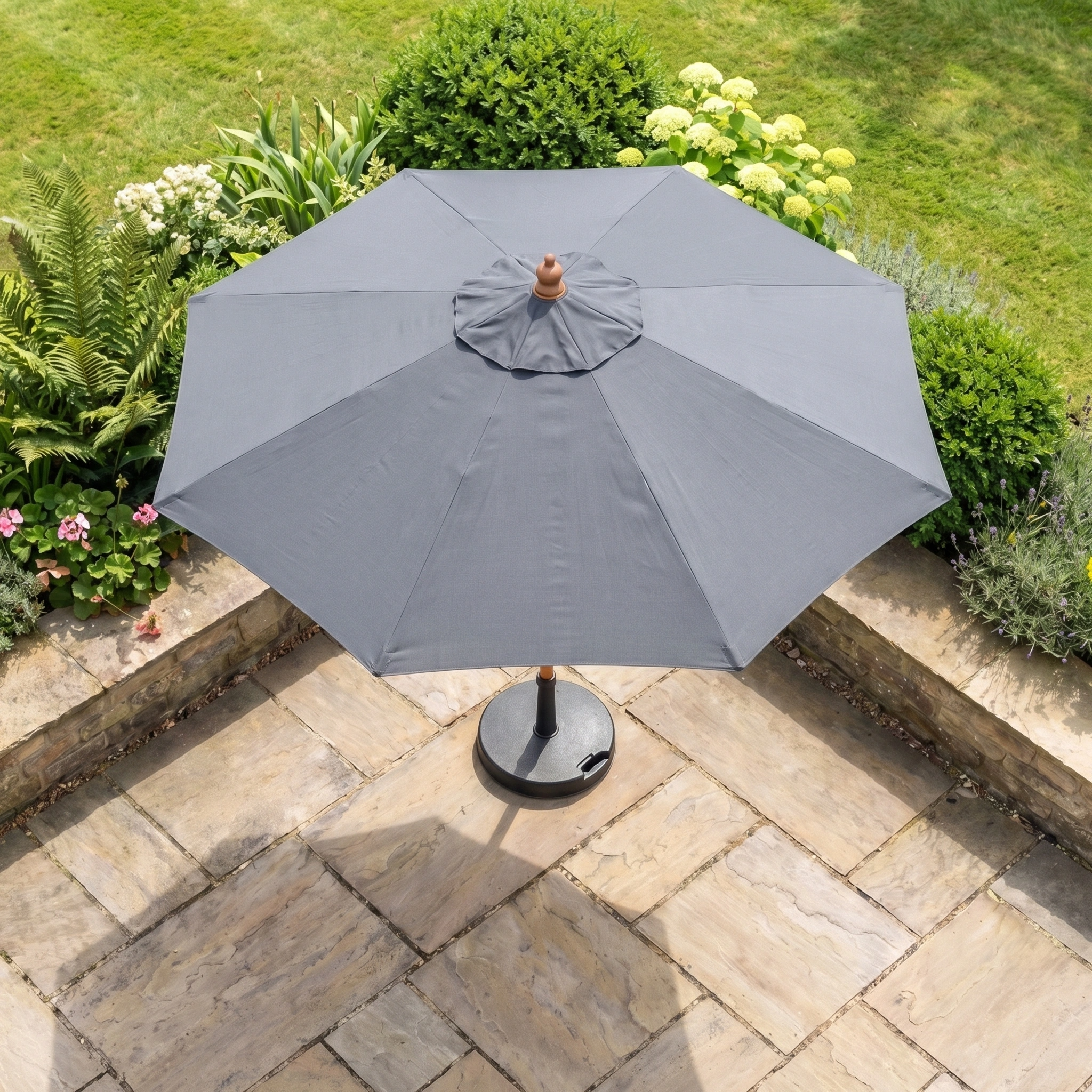 The Kratos 2m Round Wooden Parasol in Grey provides UV 50+ protection as it stands on a stone patio, surrounded by green bushes and flowering garden plants, offering reliable shade outdoors.