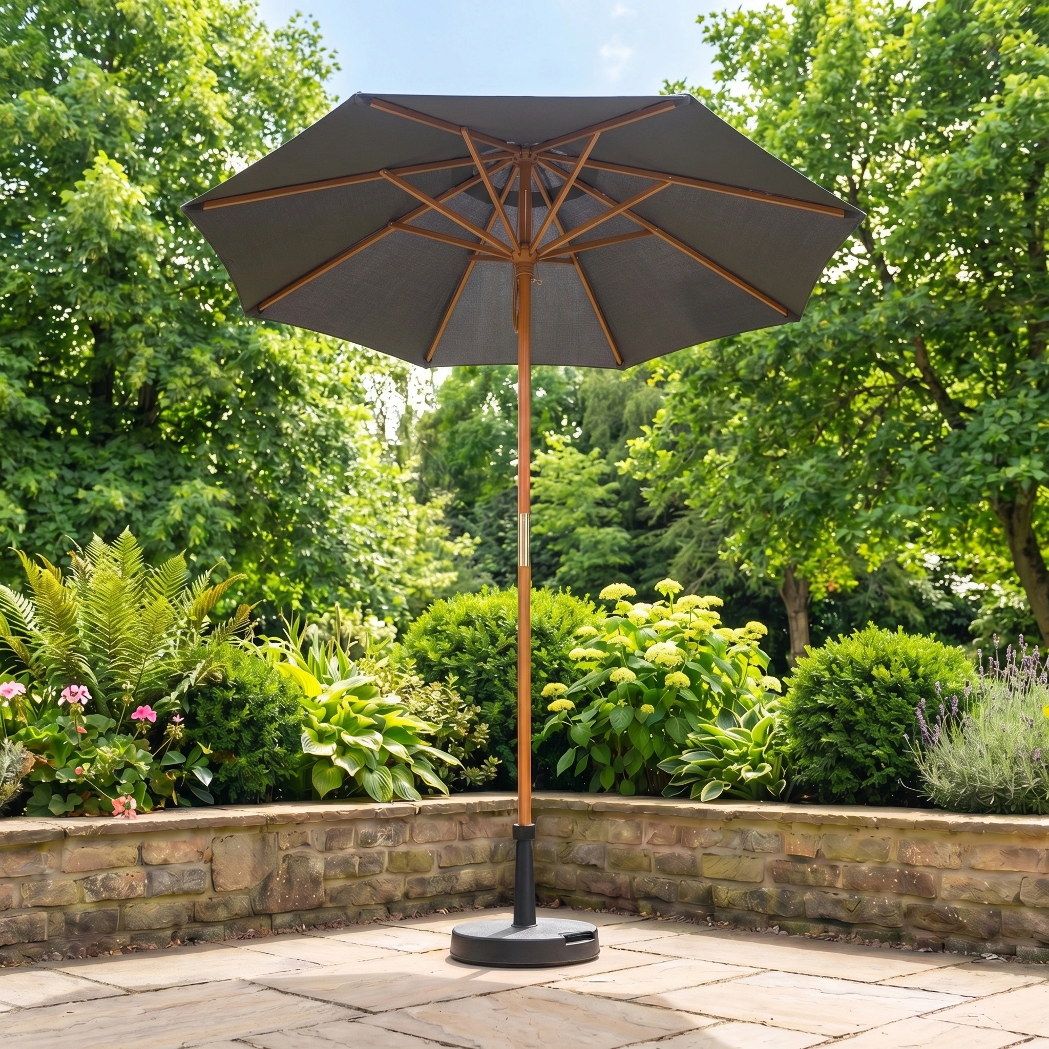 The Kratos 2m Round Wooden Parasol in Grey with UV 50+ stands on a stone terrace, surrounded by lush green plants and trees.