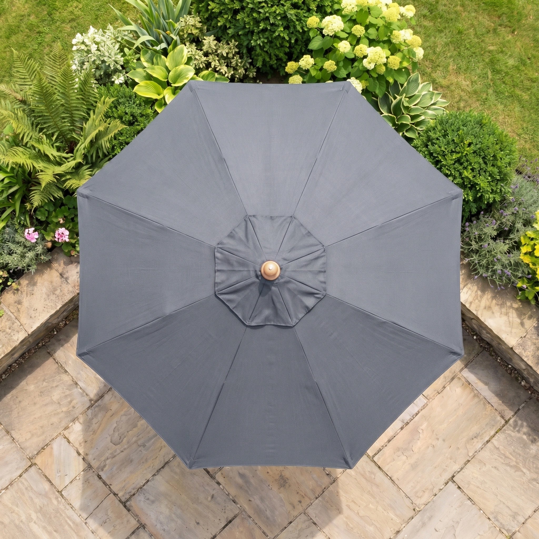 The Kratos 2m Round Wooden Parasol in Grey, offering UV50+ protection, is shown from above on a stone patio amid green garden plants.