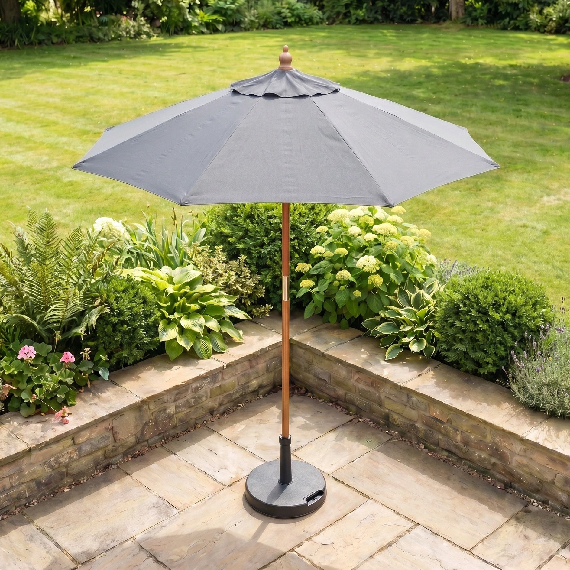 A Kratos 2m Round Wooden Parasol in Grey with UV 50+ rating stands at the center of a stone patio, surrounded by lush green plants and lawn.