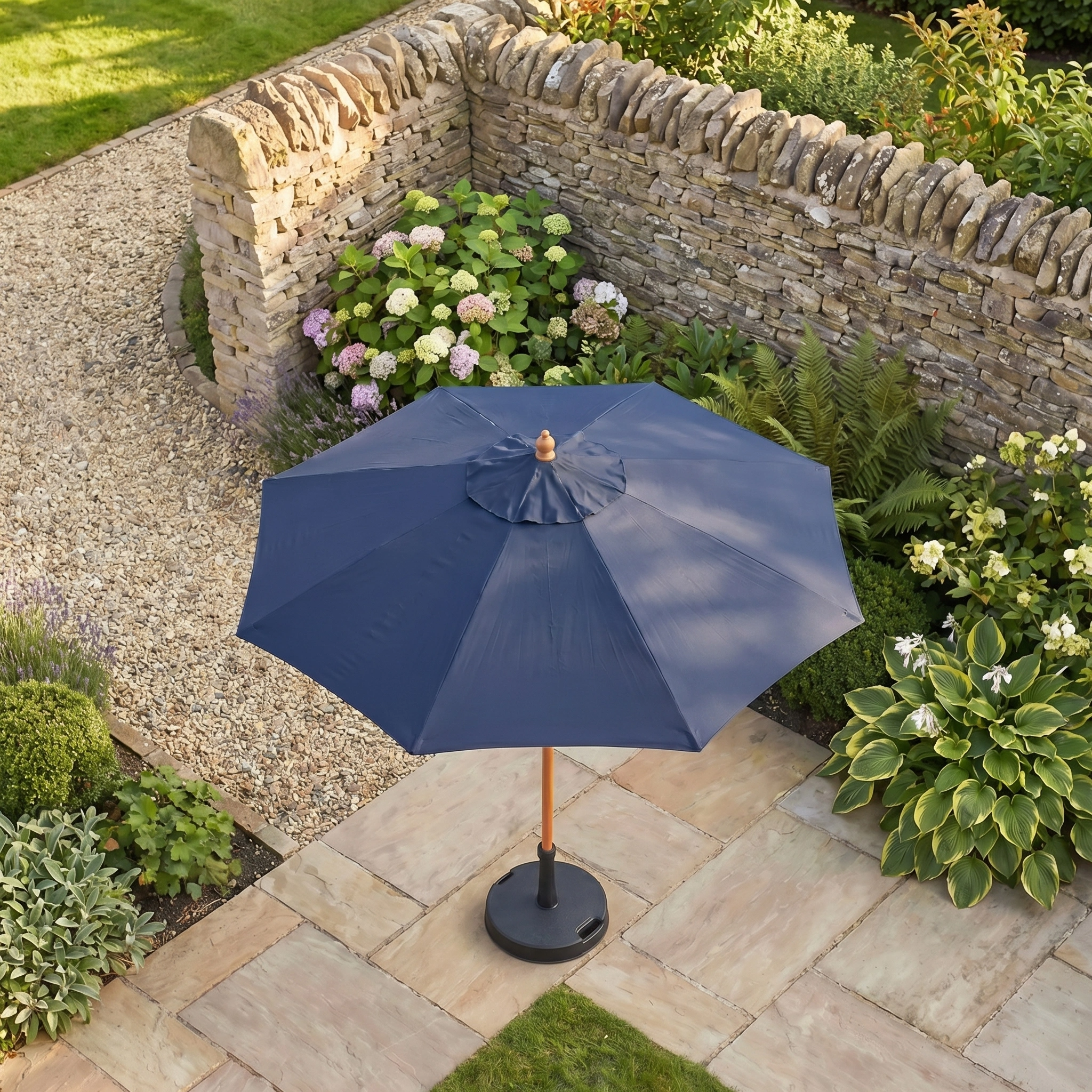 The Kratos 2m Round Wooden Parasol in Navy provides relaxing shade on stone tiles in a garden with green plants and a stone wall.