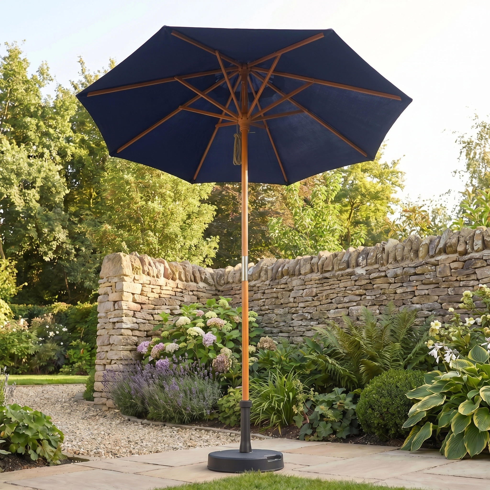 The Kratos 2m Round Wooden Parasol in Navy adds style and shade to a garden with stone walls, flowers, and green plants.