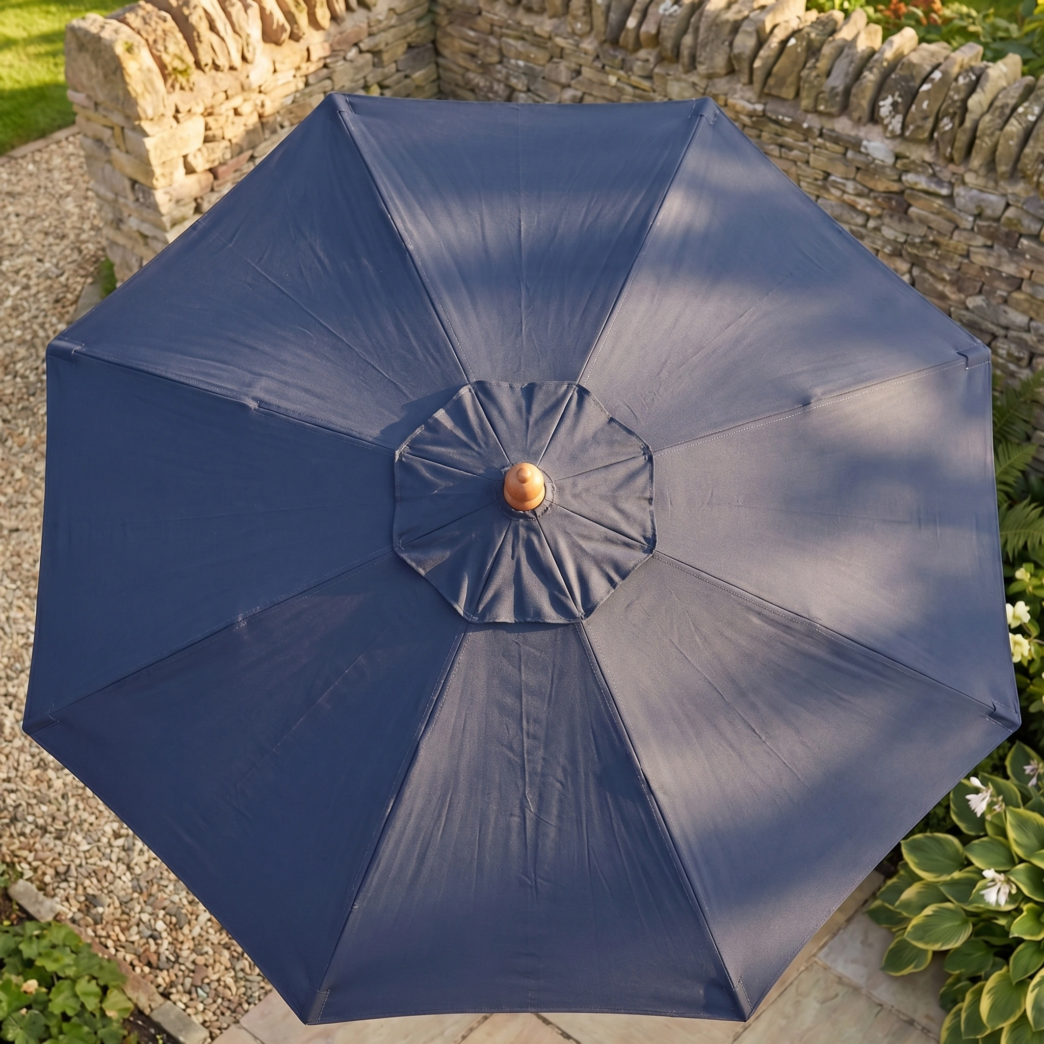 Top view of the Kratos 2m Round Wooden Parasol in Navy, open in a stone-walled garden, offering stylish shade and UV protection.