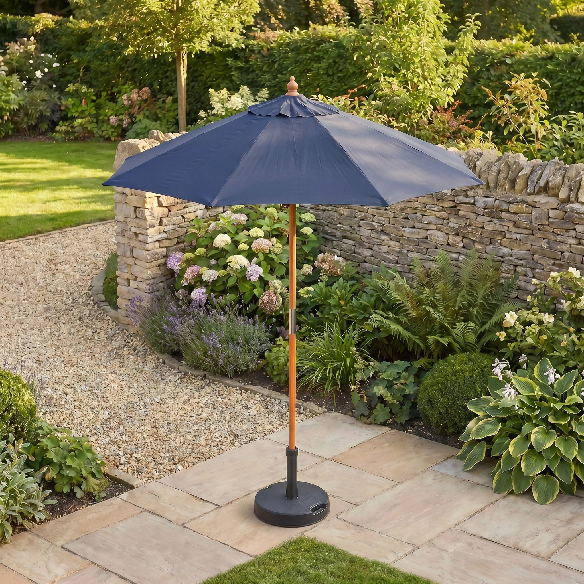 The Kratos 2m Round Wooden Parasol in Navy offers stylish shade on a stone terrace amid garden greenery and flowers.