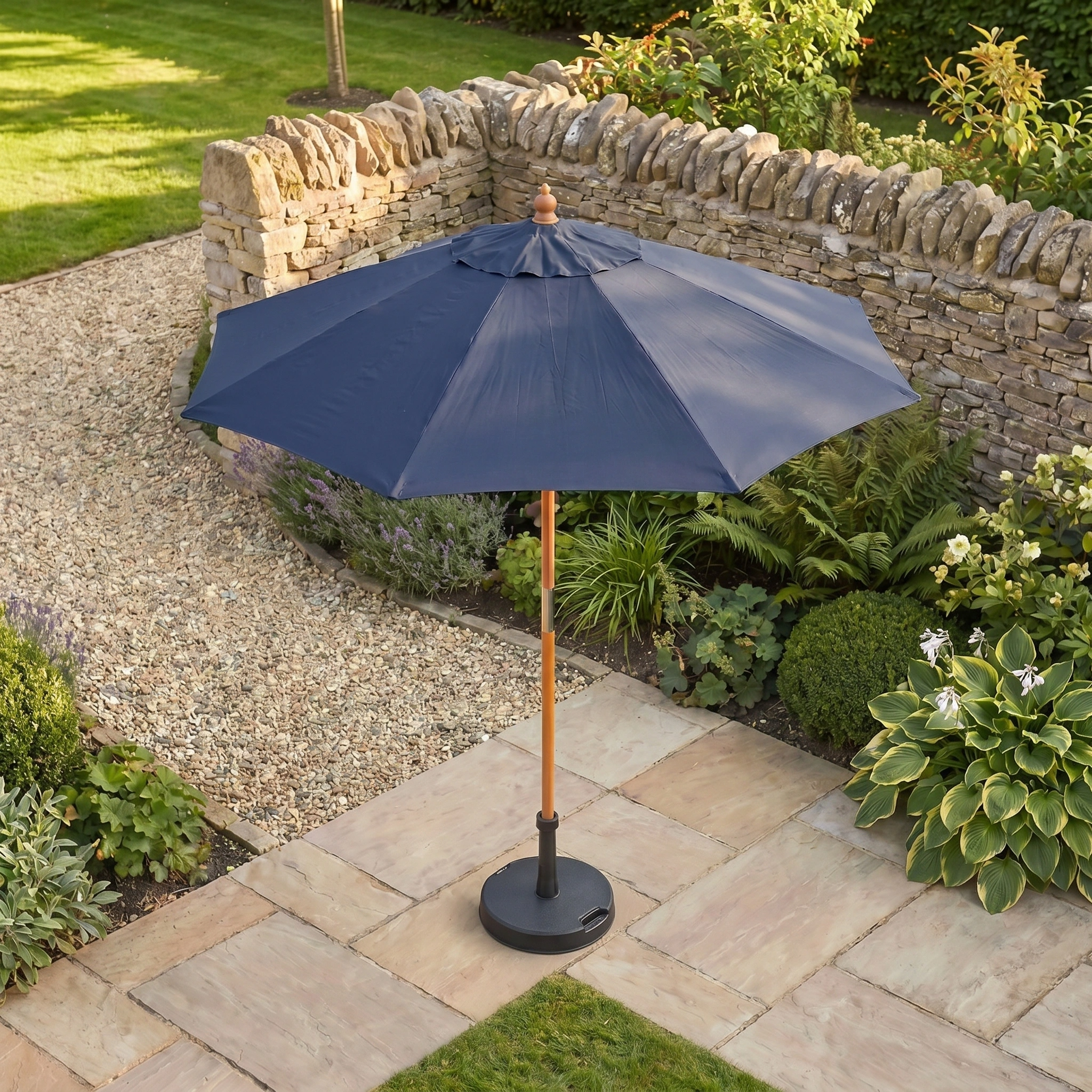 The Kratos 2m Round Wooden Parasol in Navy stands on stone tiles near a garden with plants and a stone wall.