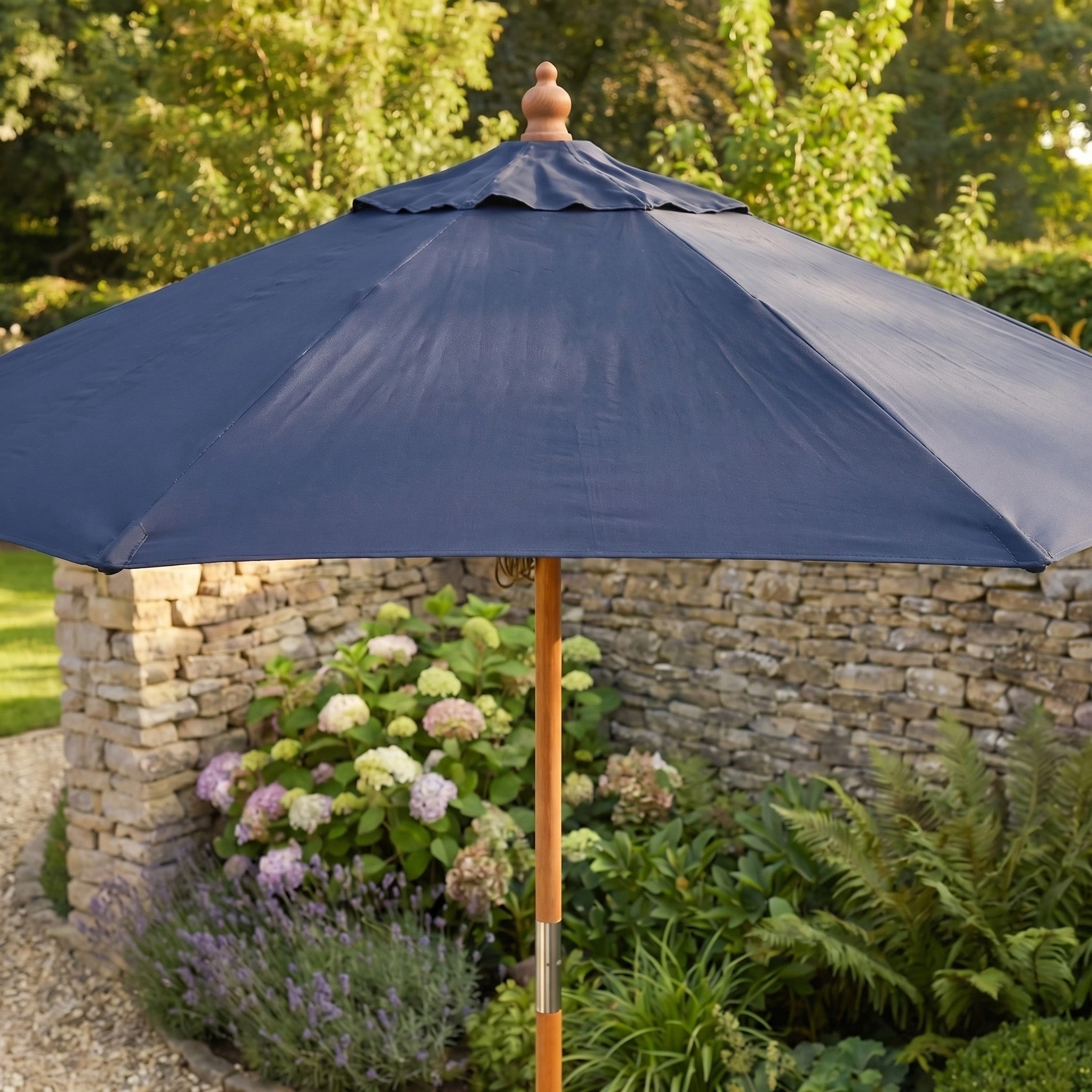 The Kratos 2m Round Wooden Parasol in Navy stands near a stone wall, surrounded by flowers and greenery, providing both elegance and shade.