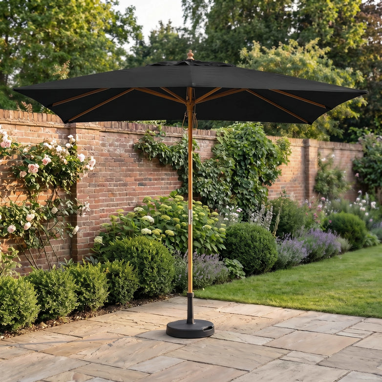 The Kratos 3m x 2m Rectangular Wooden Parasol in Black stands on a stone terrace in a garden with green bushes and a brick wall, providing stylish sun protection.