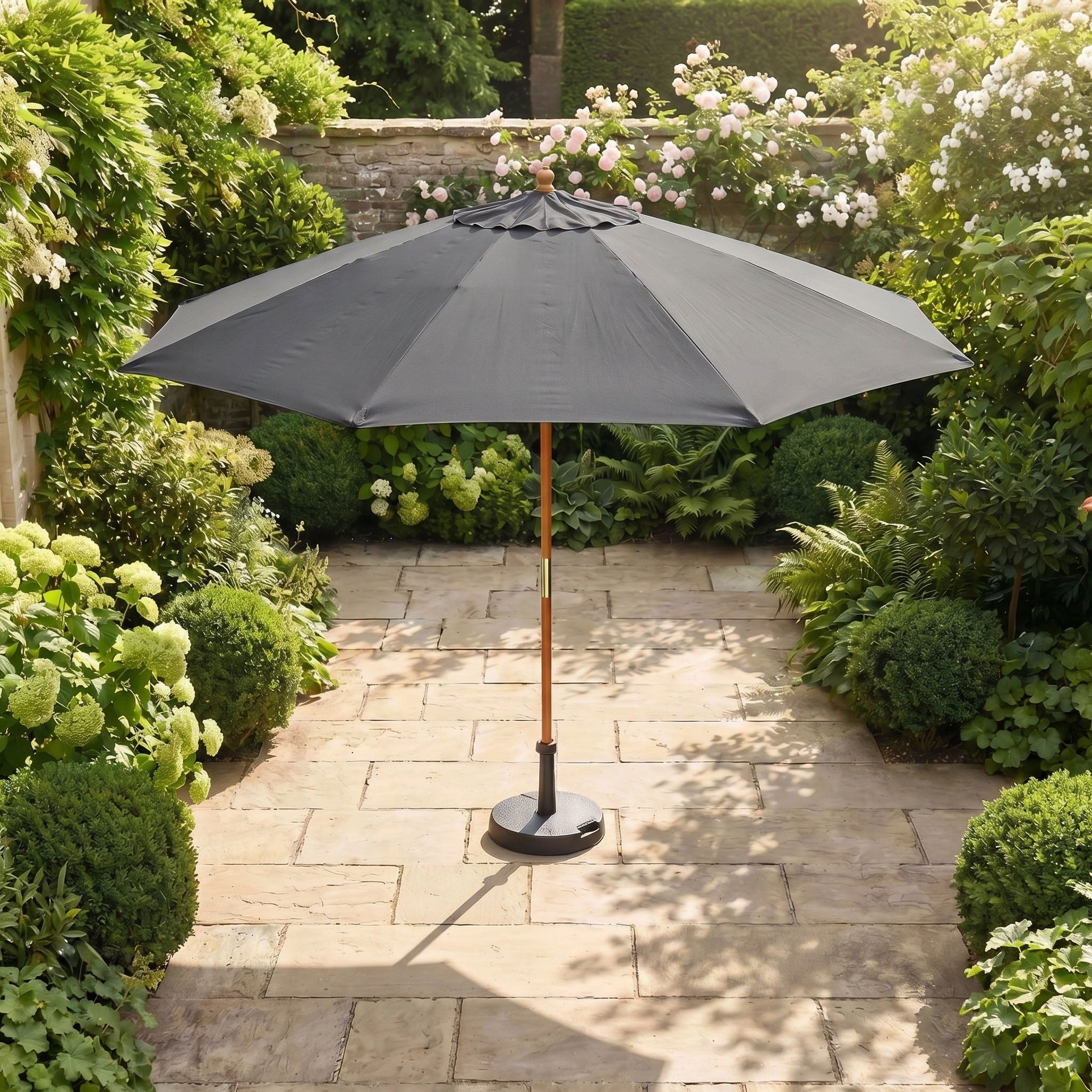 The Kratos 3m Round Wooden Parasol in Grey stands open on a stone terrace, surrounded by lush green plants and bushes, offering stylish shade and UV protection.
