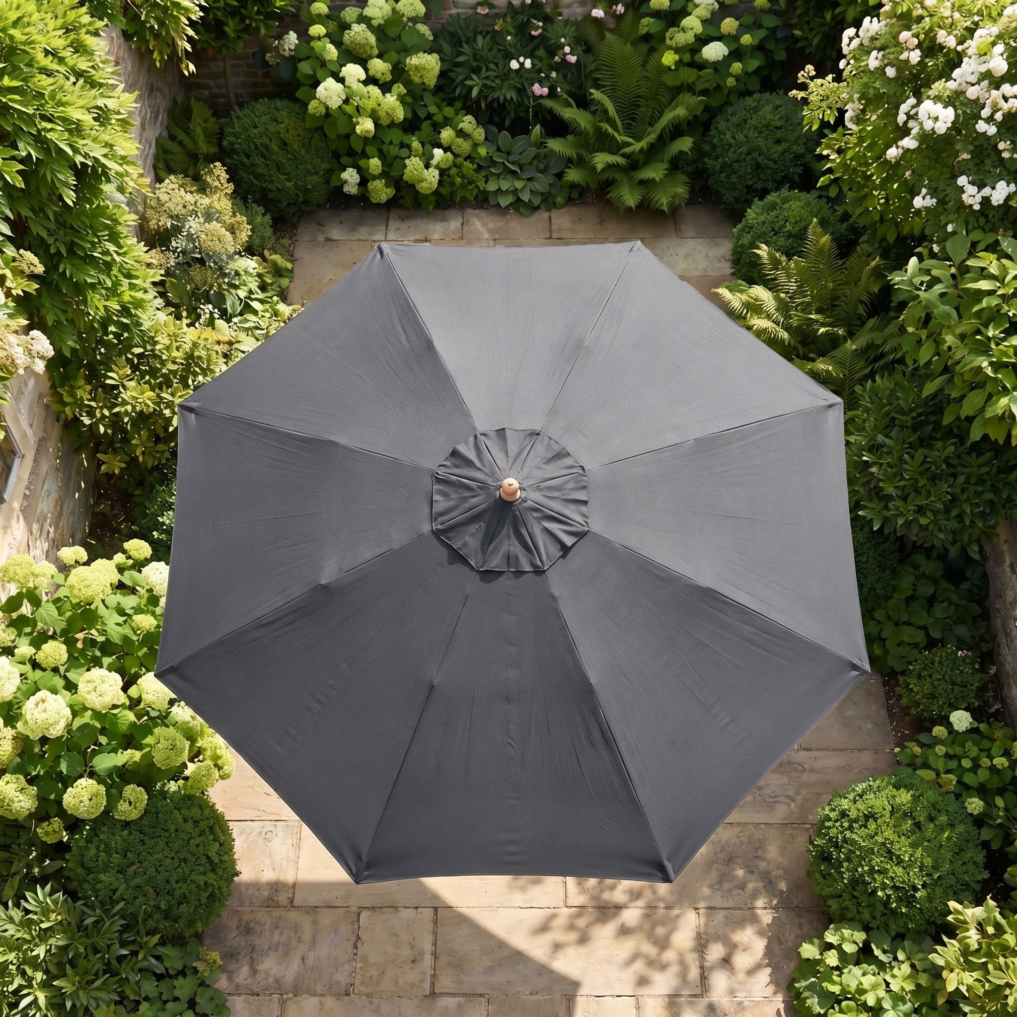 The Kratos 3m Round Wooden Parasol in Grey, viewed from above, stands out among vibrant green plants and stone paving.