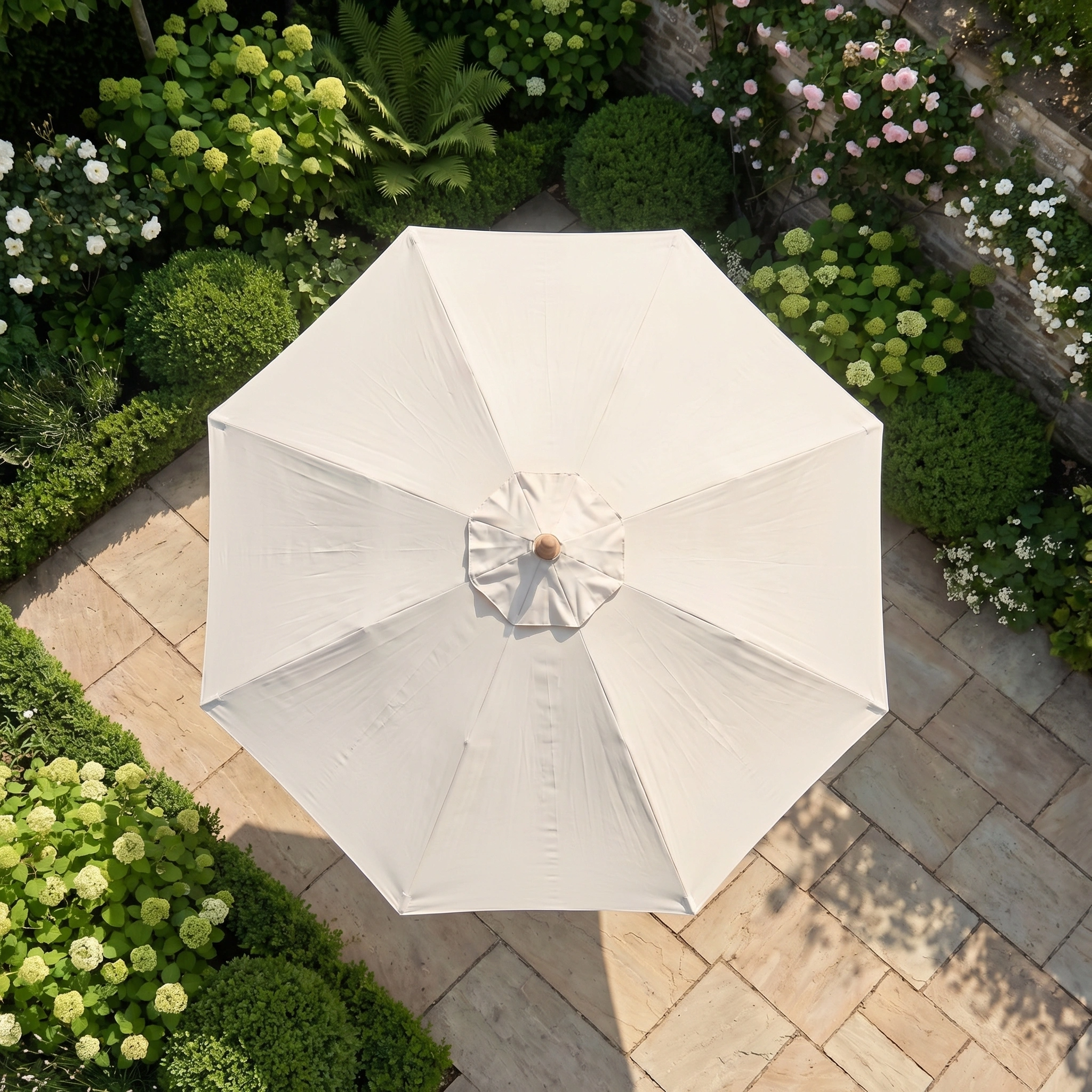 A Kratos 3m Round Wooden Parasol in Beige is seen from above, surrounded by lush green bushes and stone paving in a garden, offering stylish UV protection.