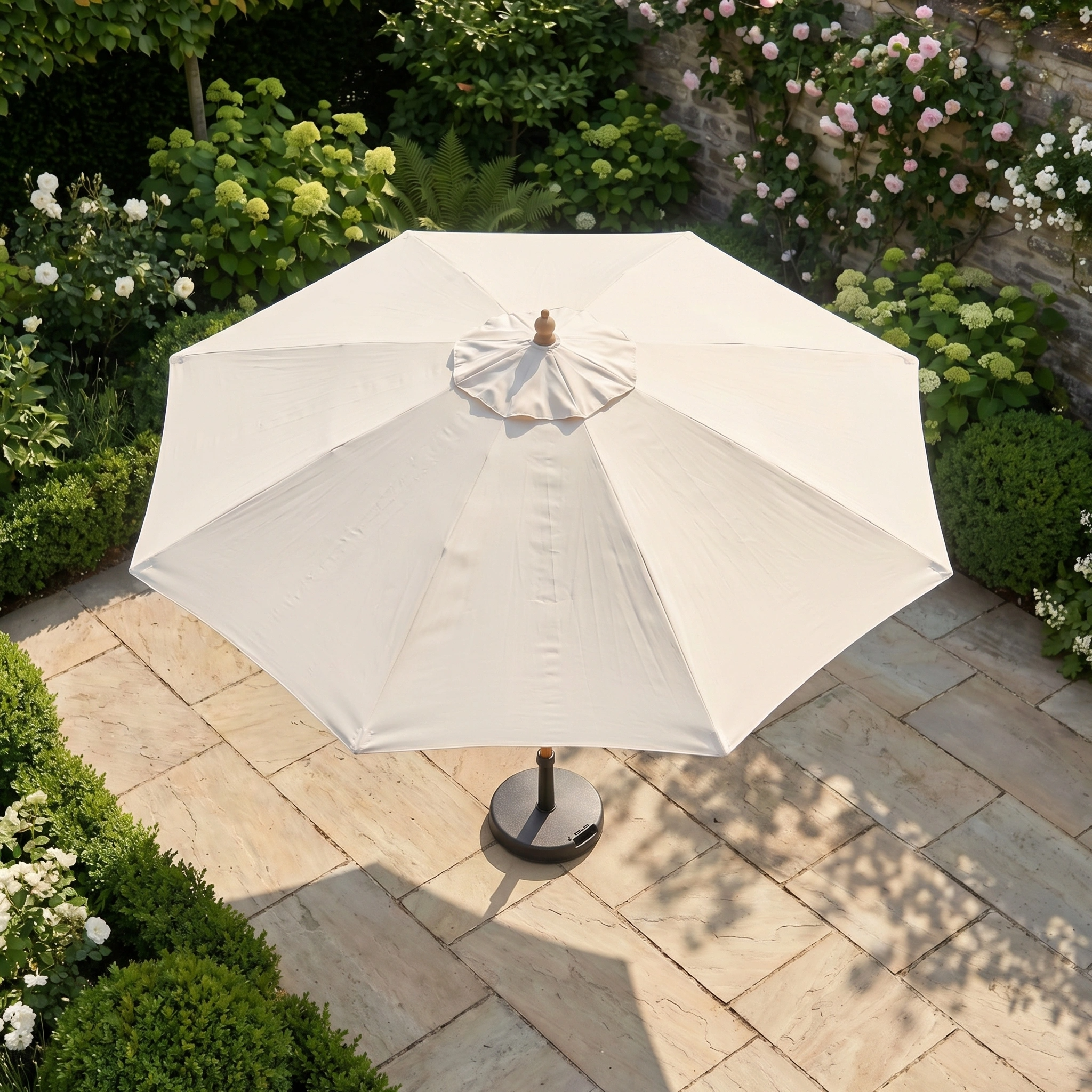 The Kratos 3m Round Wooden Parasol in Beige provides stylish shade and UV protection on a stone patio, surrounded by green bushes and blooming flowers in your garden.