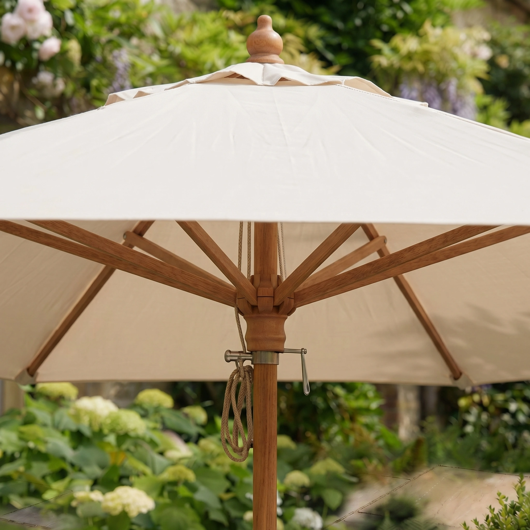 The Kratos 3m Round Wooden Parasol in Beige stands in a lush garden among green plants, providing stylish sun protection with its wooden pole and elegant design.