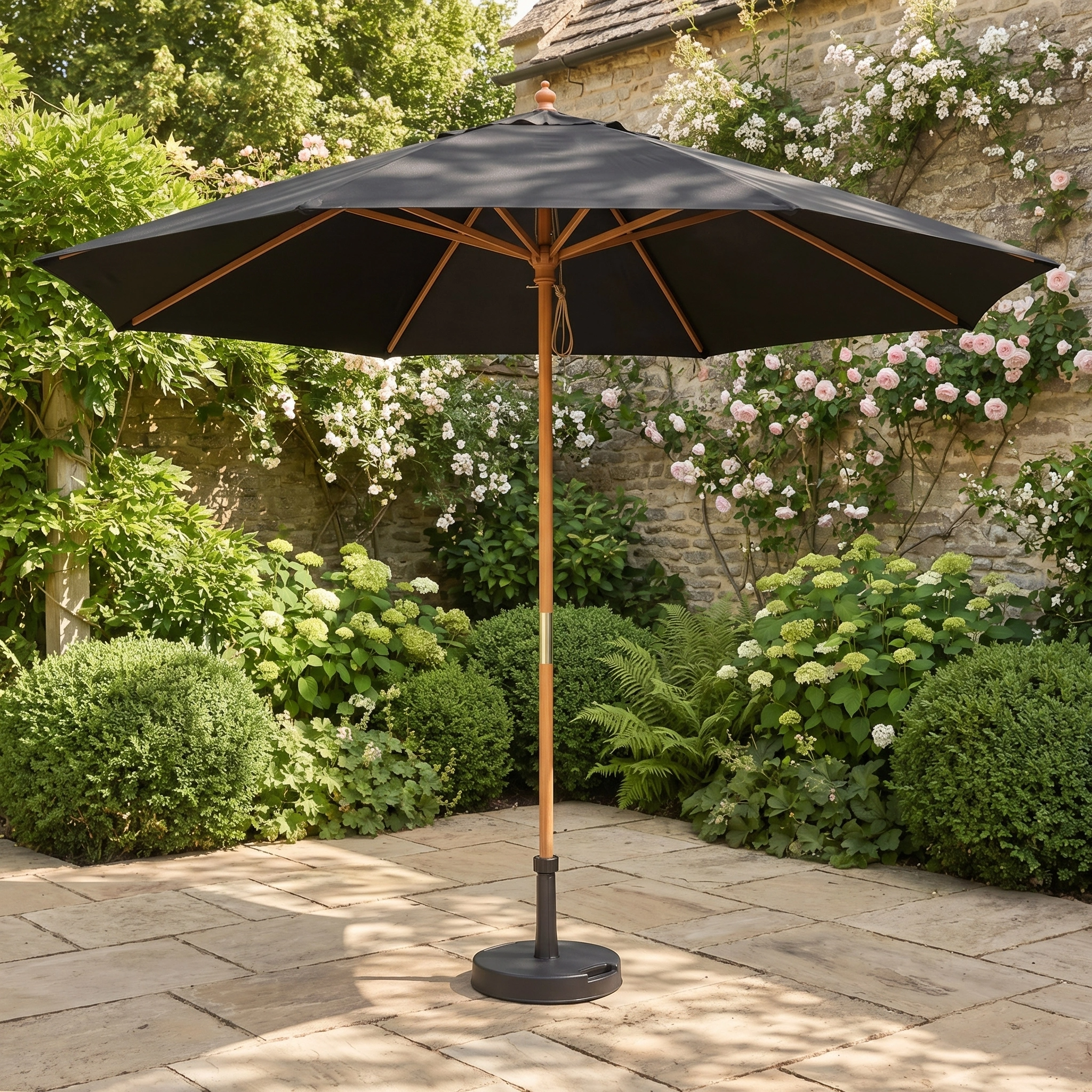 The Kratos 3m Round Wooden Parasol in Black stands on a stone terrace amid green bushes and blooming flowers, providing stylish UV sun protection for your garden.