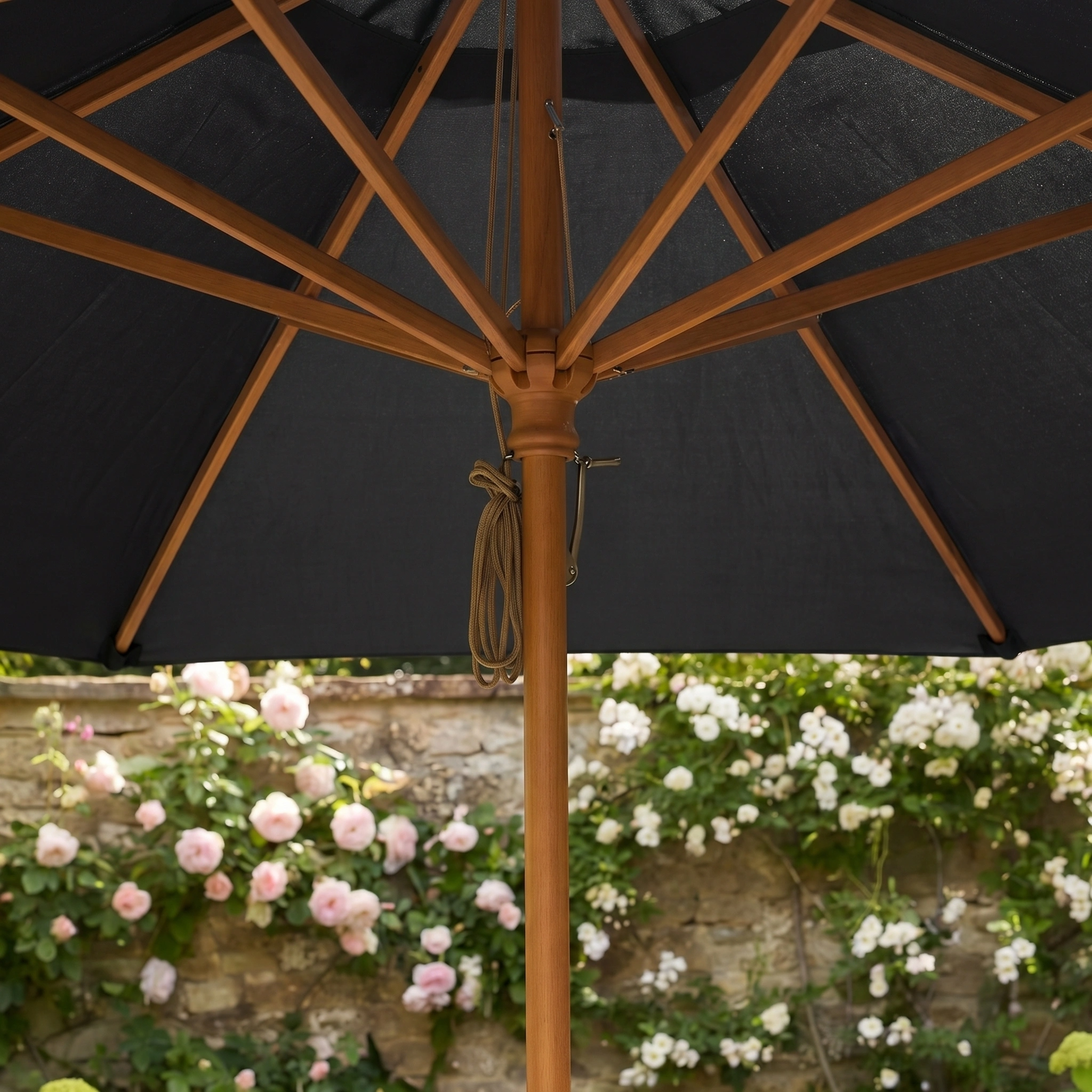 View from under the Kratos 3m Round Wooden Parasol in Black, with its UV-protective black canopy and wooden frame, set against flowering bushes and a stone wall backdrop.