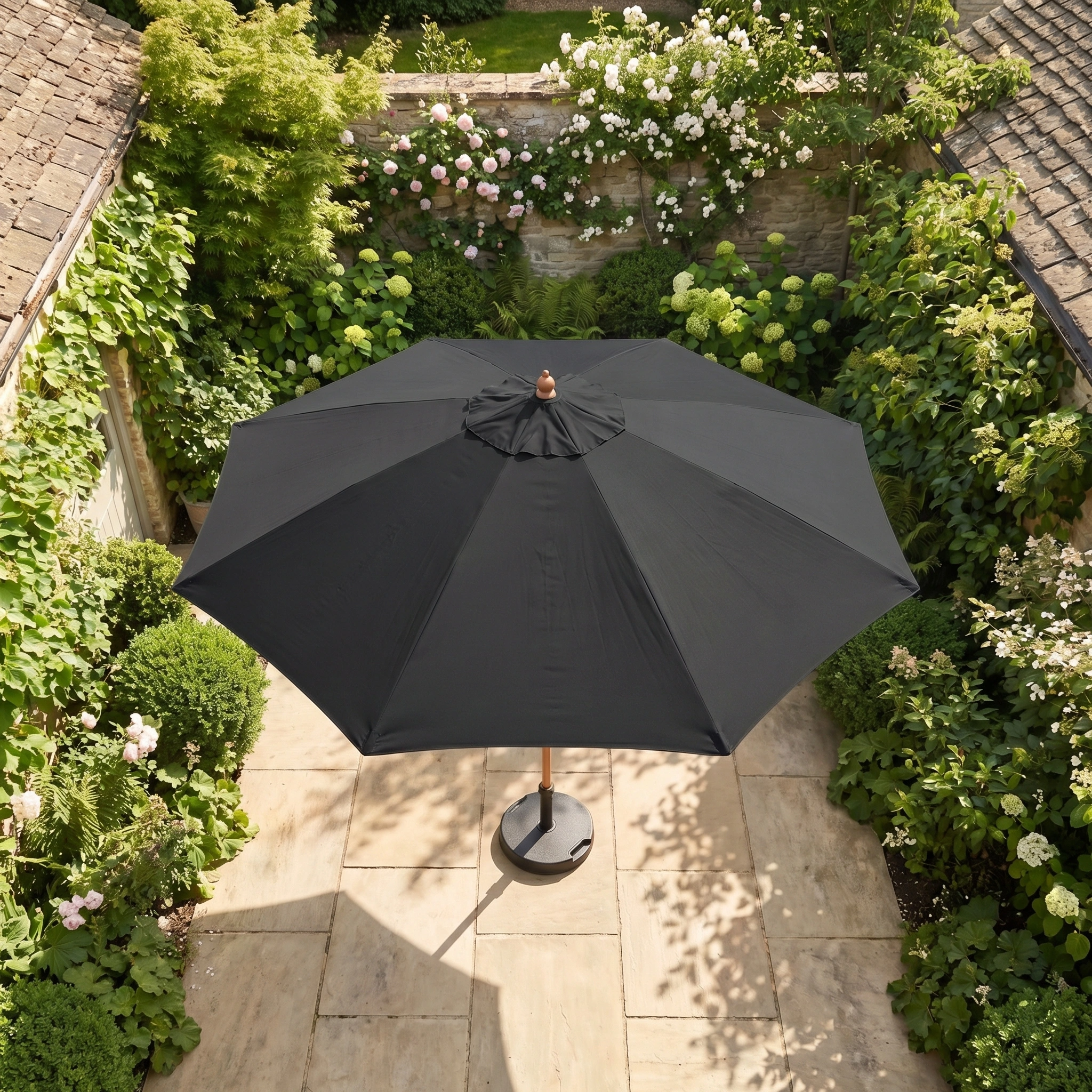 The Kratos 3m Round Wooden Parasol in Black provides UV sun protection, perfect for relaxing outdoors on a stone terrace surrounded by green plants and flowering bushes.