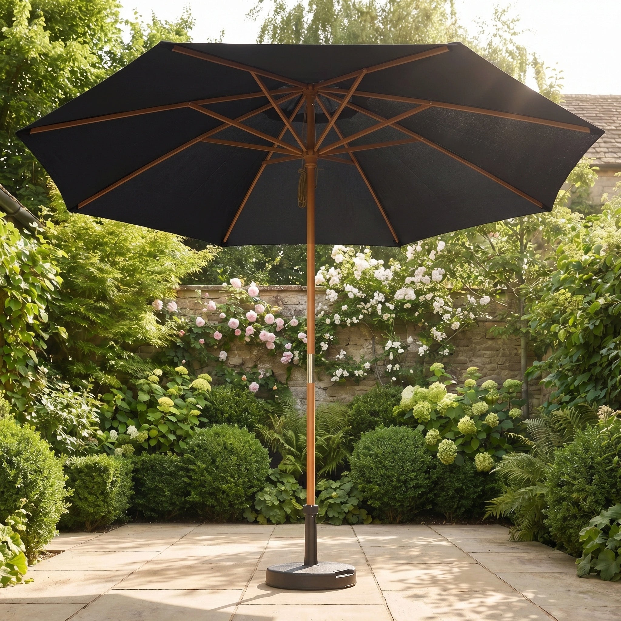 The Kratos 3m Round Wooden Parasol in Black provides UV protection as it stands open on a sunny stone terrace, surrounded by lush green garden plants.