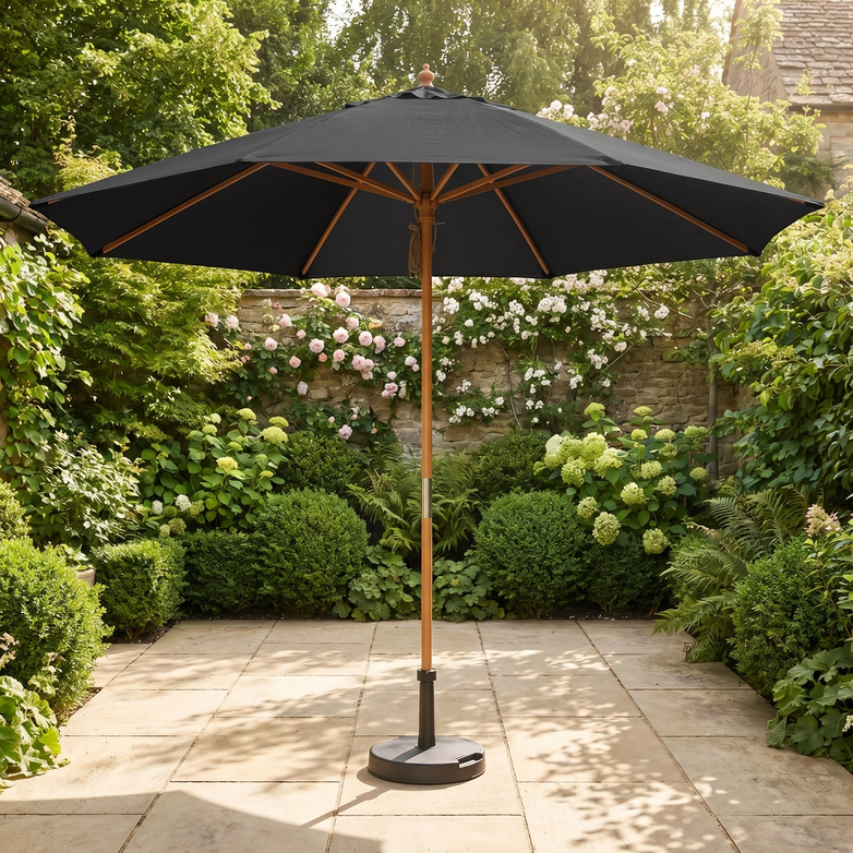 The Kratos 3m Round Wooden Parasol in Black stands on a stone terrace, providing UV sun protection among lush green bushes and blooming flowers.