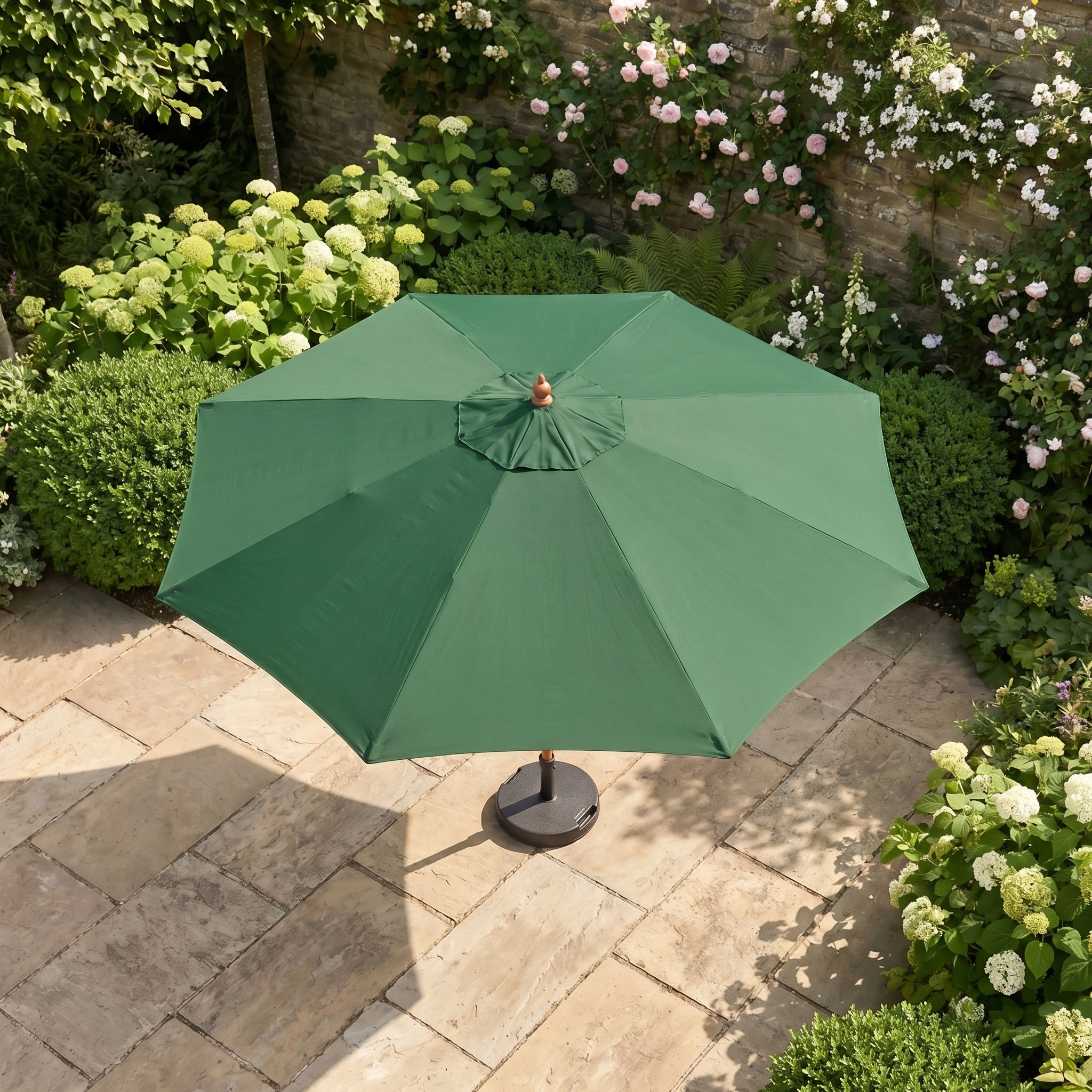 The Kratos 3m Round Wooden Parasol in Green shades your stone patio, surrounded by lush bushes and flowering plants. Enjoy outdoor comfort and style with this UV-protective parasol, perfect for relaxing al fresco.