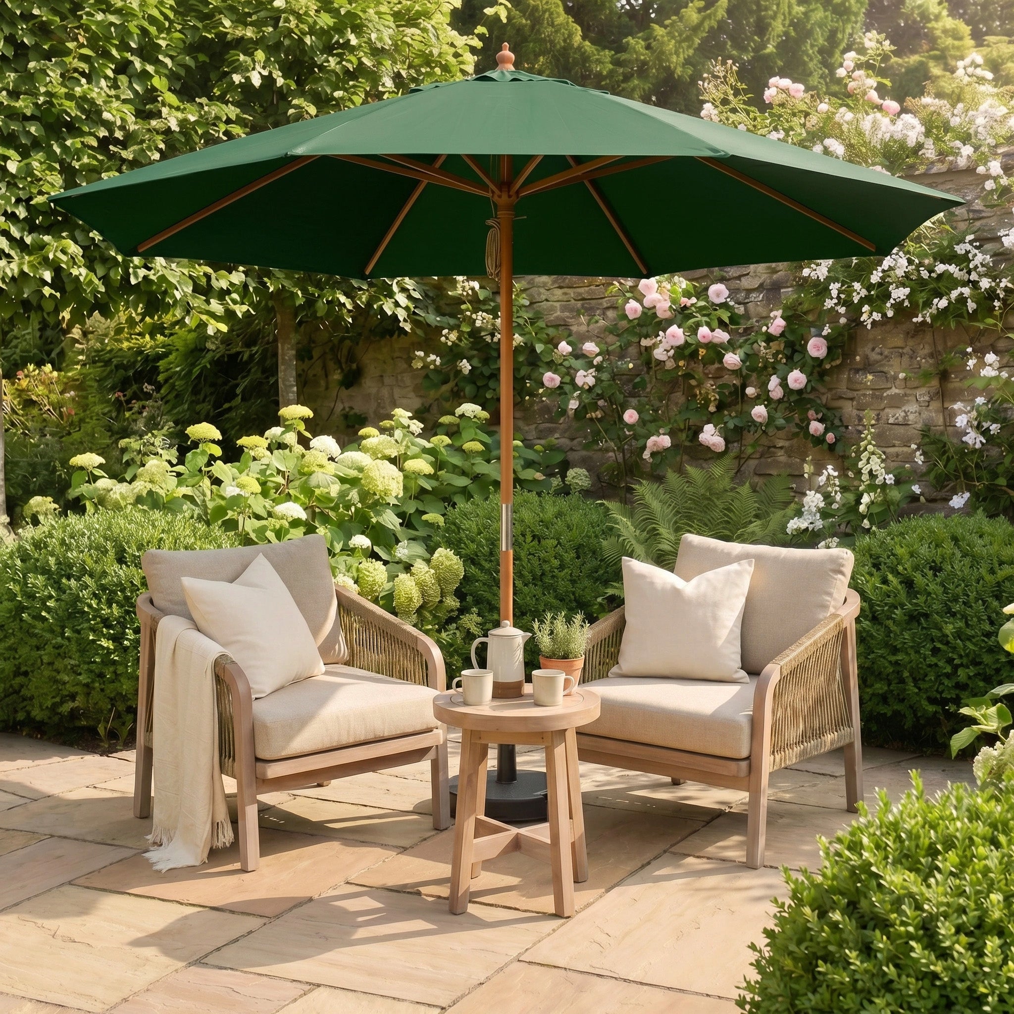 Two cushioned chairs and a table with mugs sit under the Kratos 3m Round Wooden Parasol in Green, offering UV protection in a garden filled with blooming flowers.