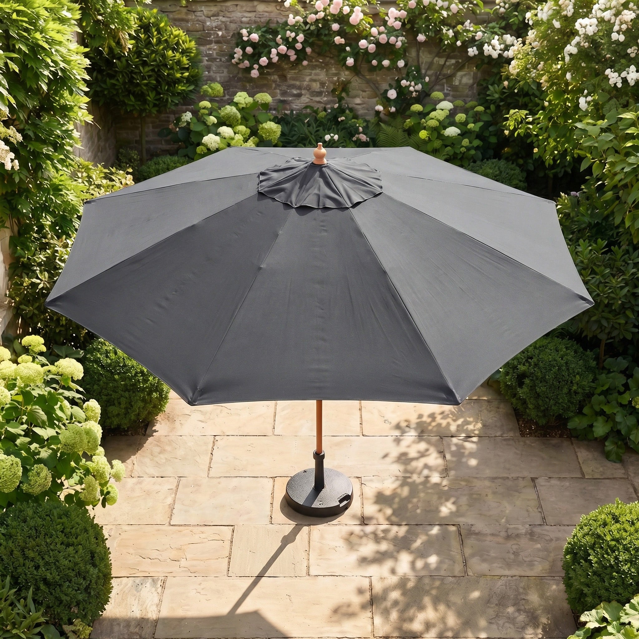 The Kratos 3m Round Wooden Parasol in Grey stands on stone tiles in a lush garden surrounded by green bushes and blooming flowers.