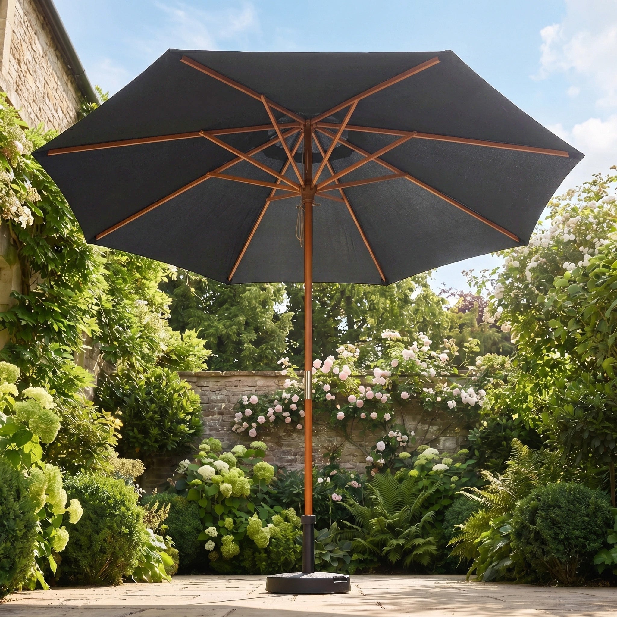 The Kratos 3m Round Wooden Parasol in Grey stands on a stone terrace, its large grey canopy providing UV protection among lush green plants and blooming flowers.