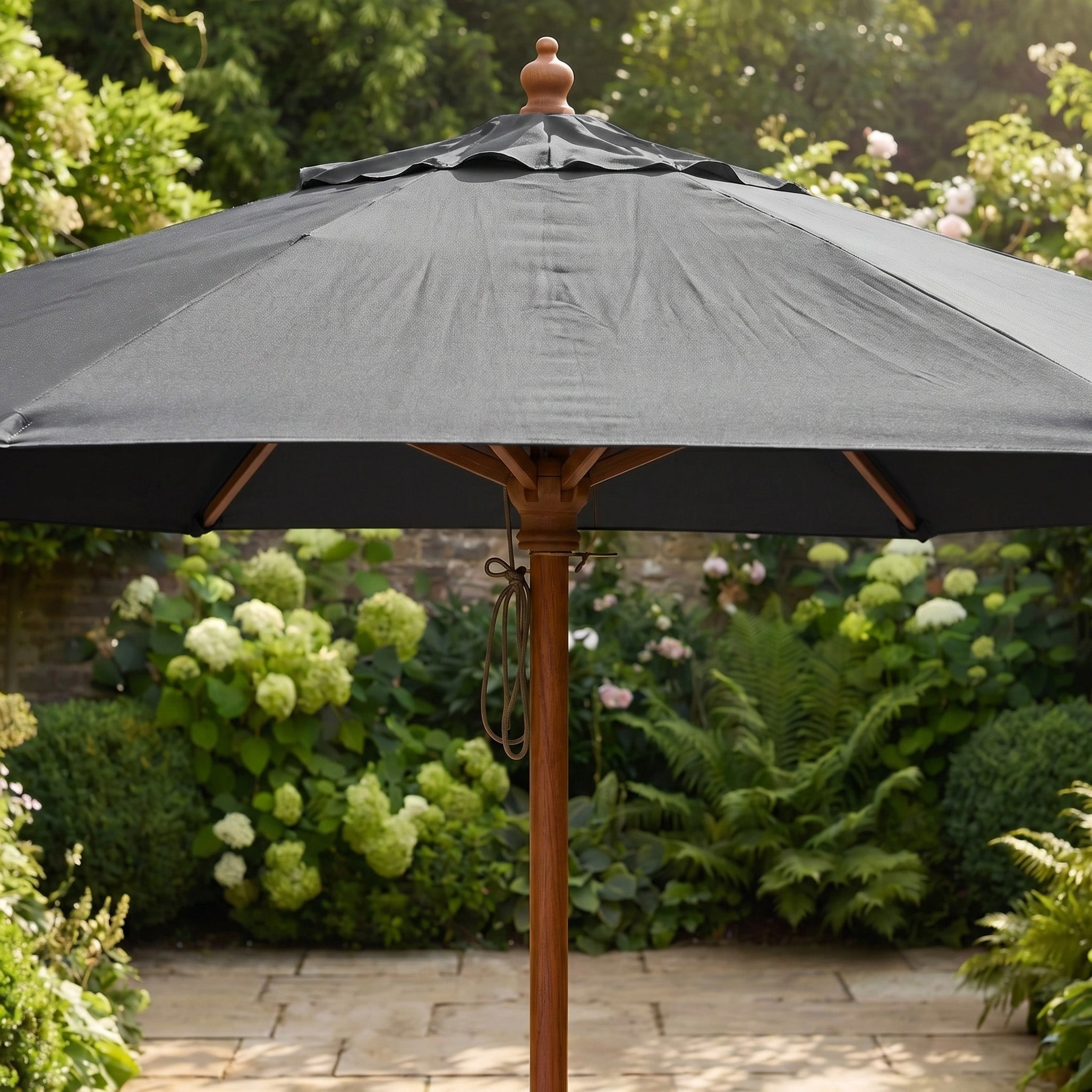 The Kratos 3m Round Wooden Parasol in Grey stands over a garden patio with lush green plants and flowers in the background, offering stylish UV protection.