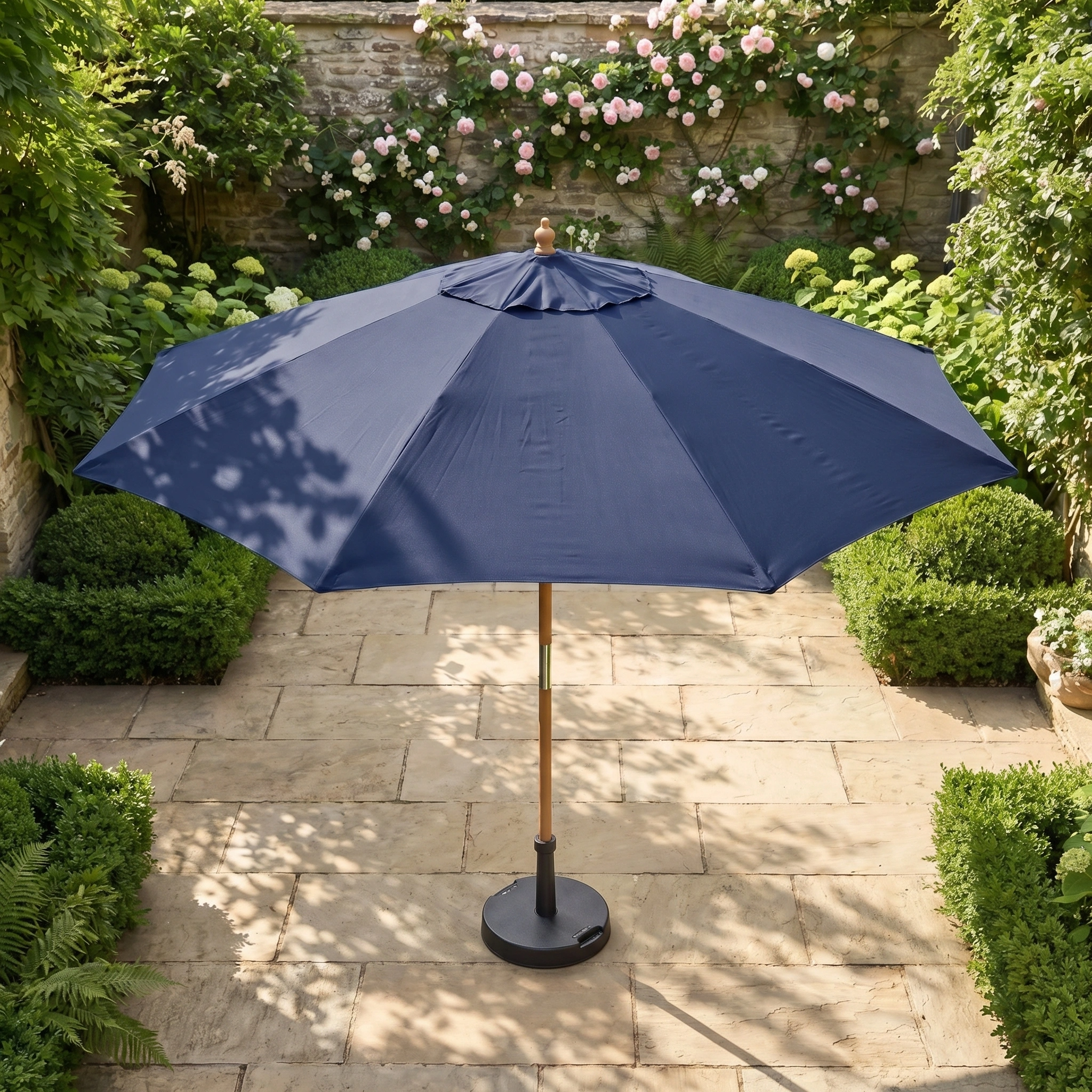 Kratos 3m Round Wooden Parasol in Navy with UV 50+ protection on a stone terrace, surrounded by green bushes and flowering plants.