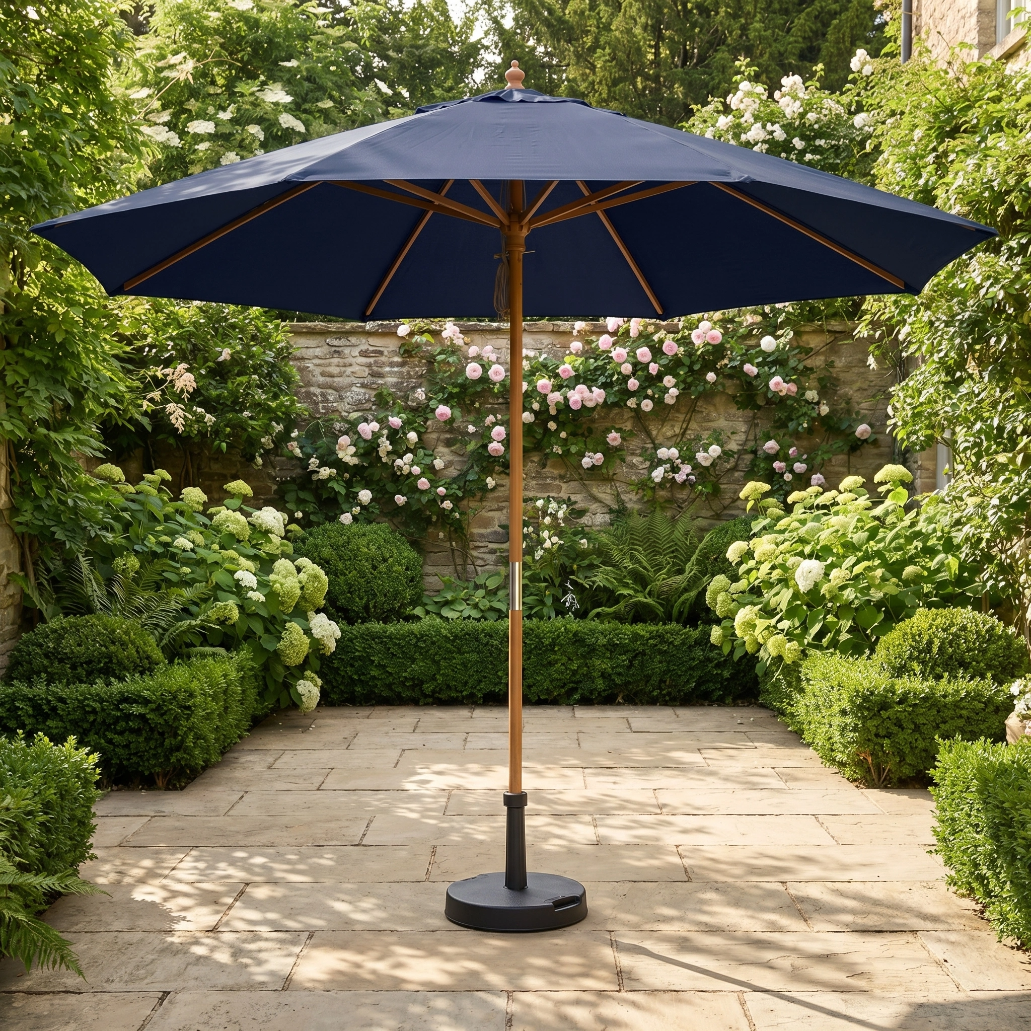 The Kratos 3m Round Wooden Parasol in Navy with UV 50+ sun protection stands on a stone terrace, surrounded by lush green plants and blooming flowers.